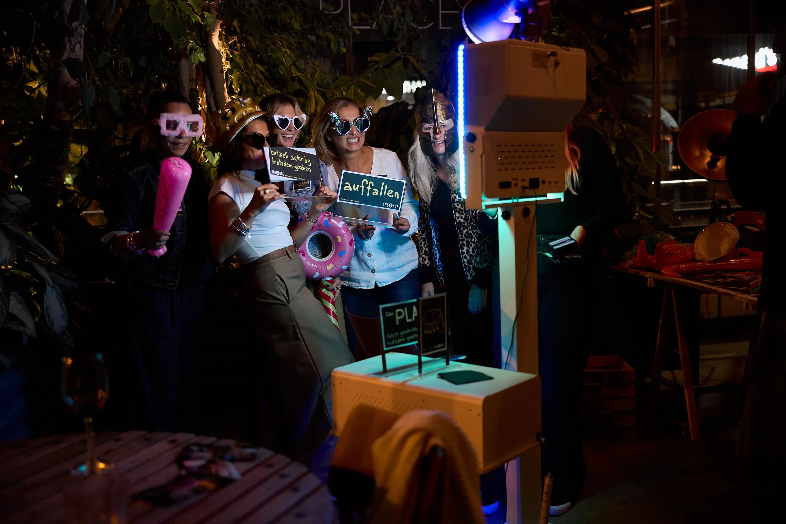 Gruppe junger Frauen mit Party-Accessoires vor Fotostation mit Kamera, bei einer Party im Außenbereich bei Nacht, mit bunten Lichtern und Dekorationen.