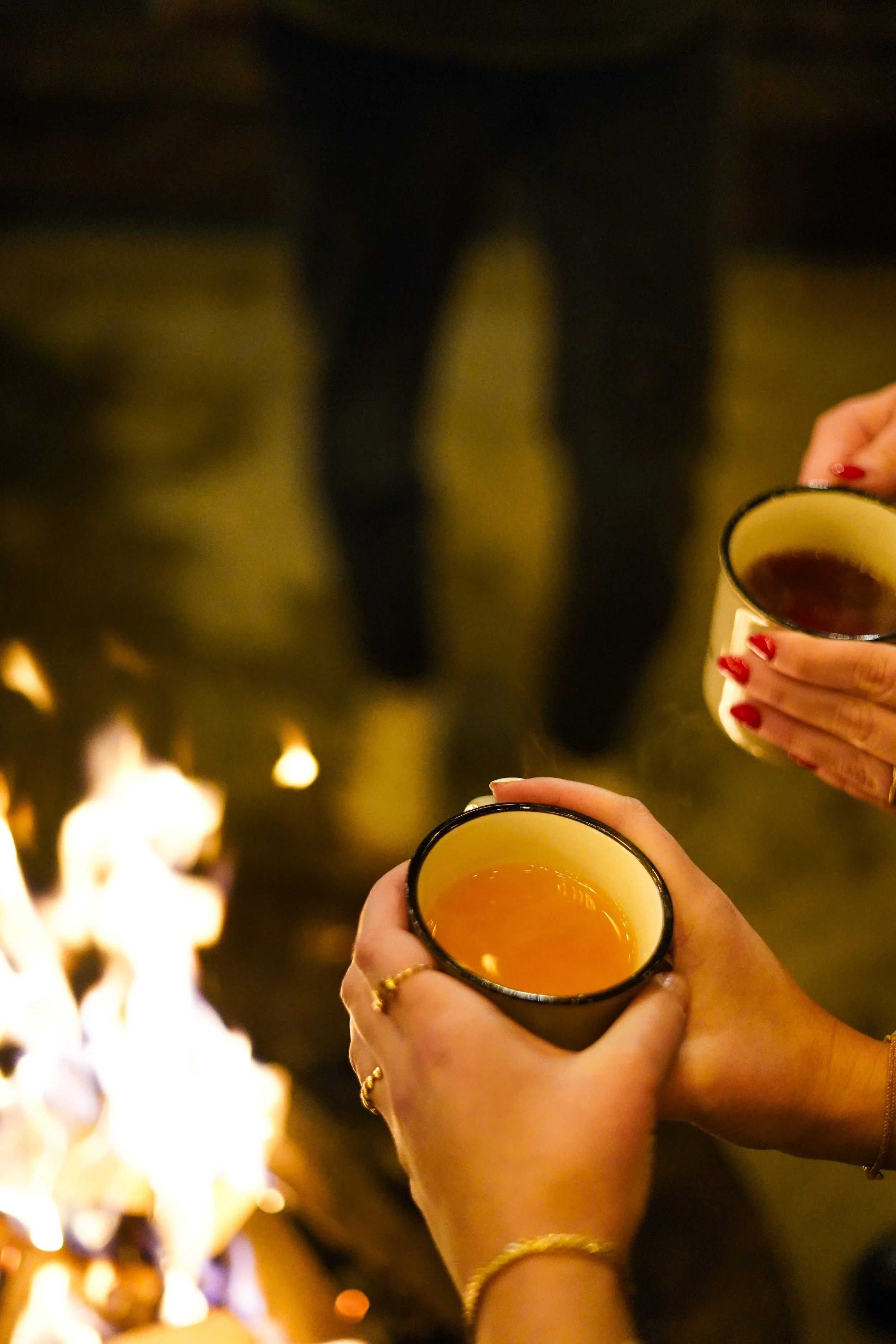 Zwei Frauen halten Tassen mit Heißgetränken in der Hand, während im Vordergrund ein Feuer brennt und im Hintergrund eine Person steht.