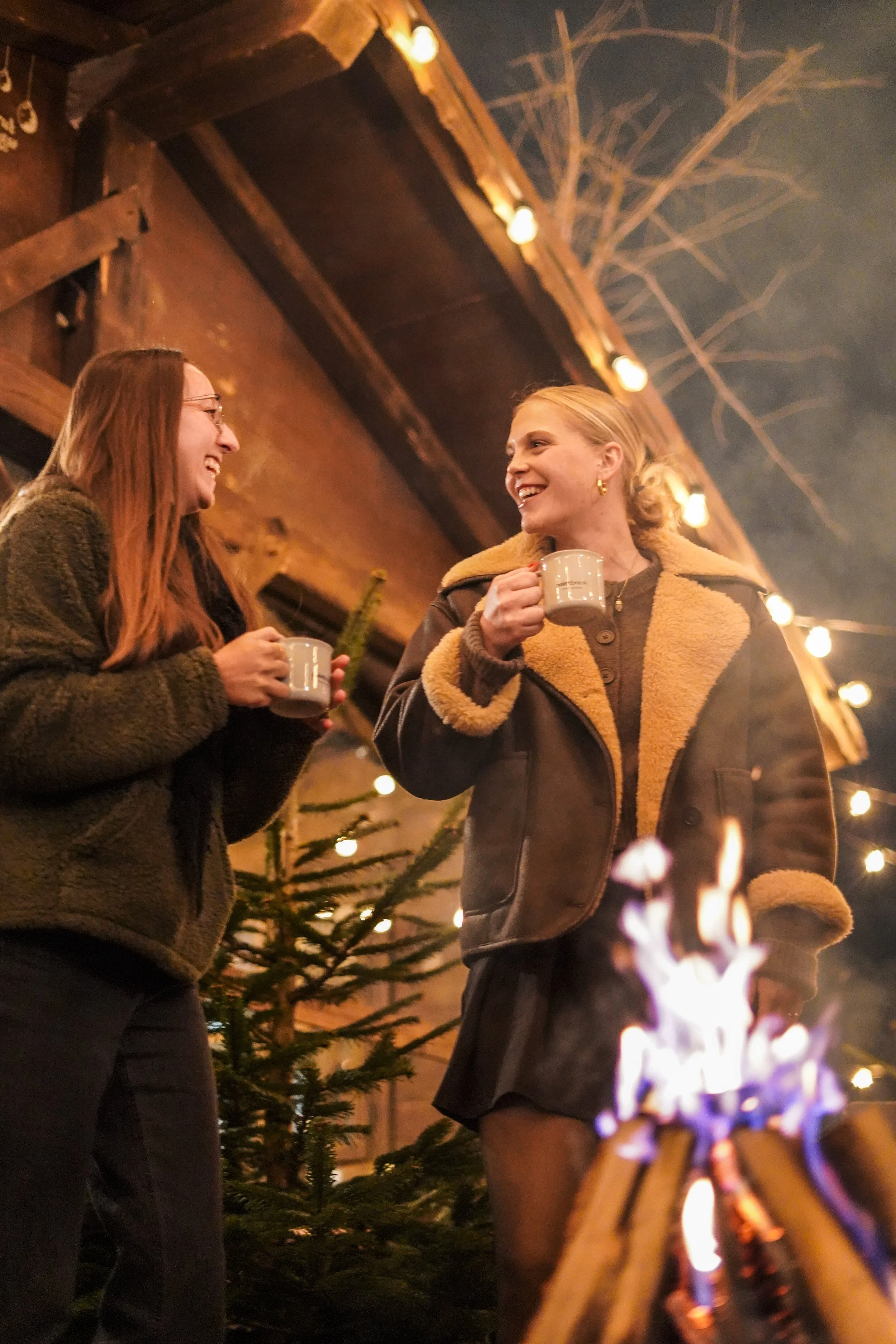 Zwei Frauen stehen beim Lagerfeuer im Freien an einem Weihnachtsbaum und unter Lichterketten, die beim Feiern im Winterabend in Weihnachtsstimmung setzen, und trinken Kaffee oder Tee.