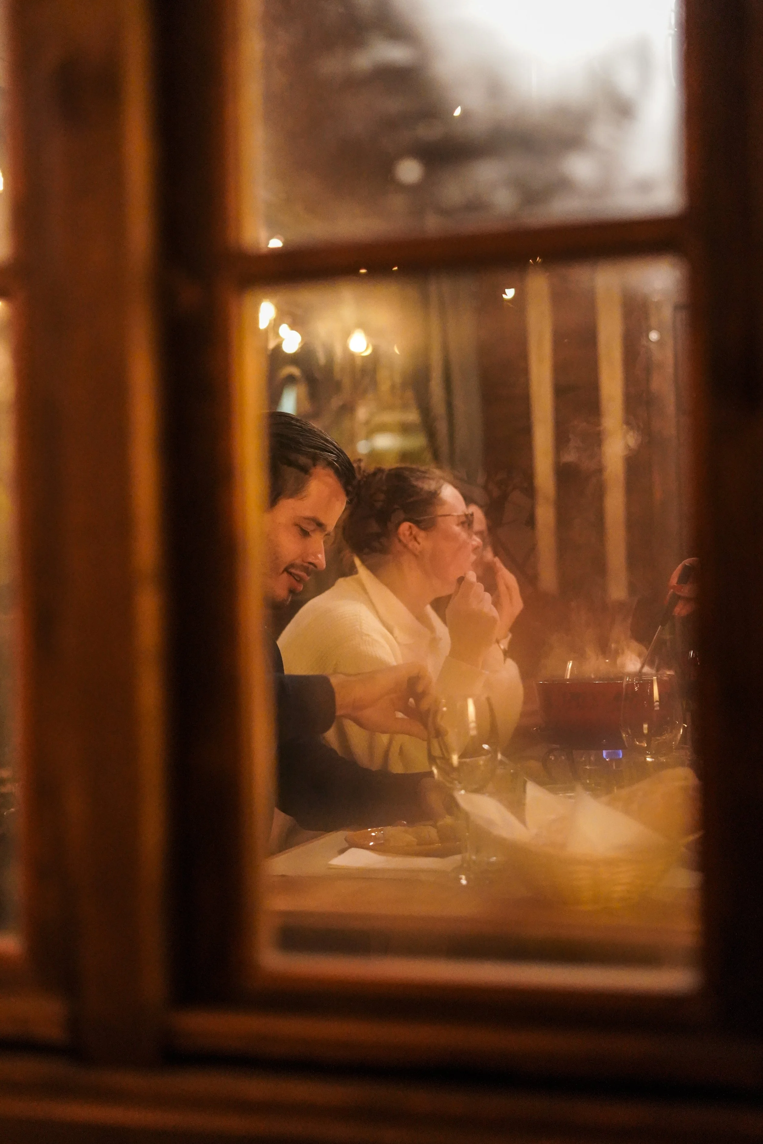 Menschen sitzen an einem Tisch in einem Ambiente mit warmem Licht, gesehen durch ein Fenster mit Holzrahmen.