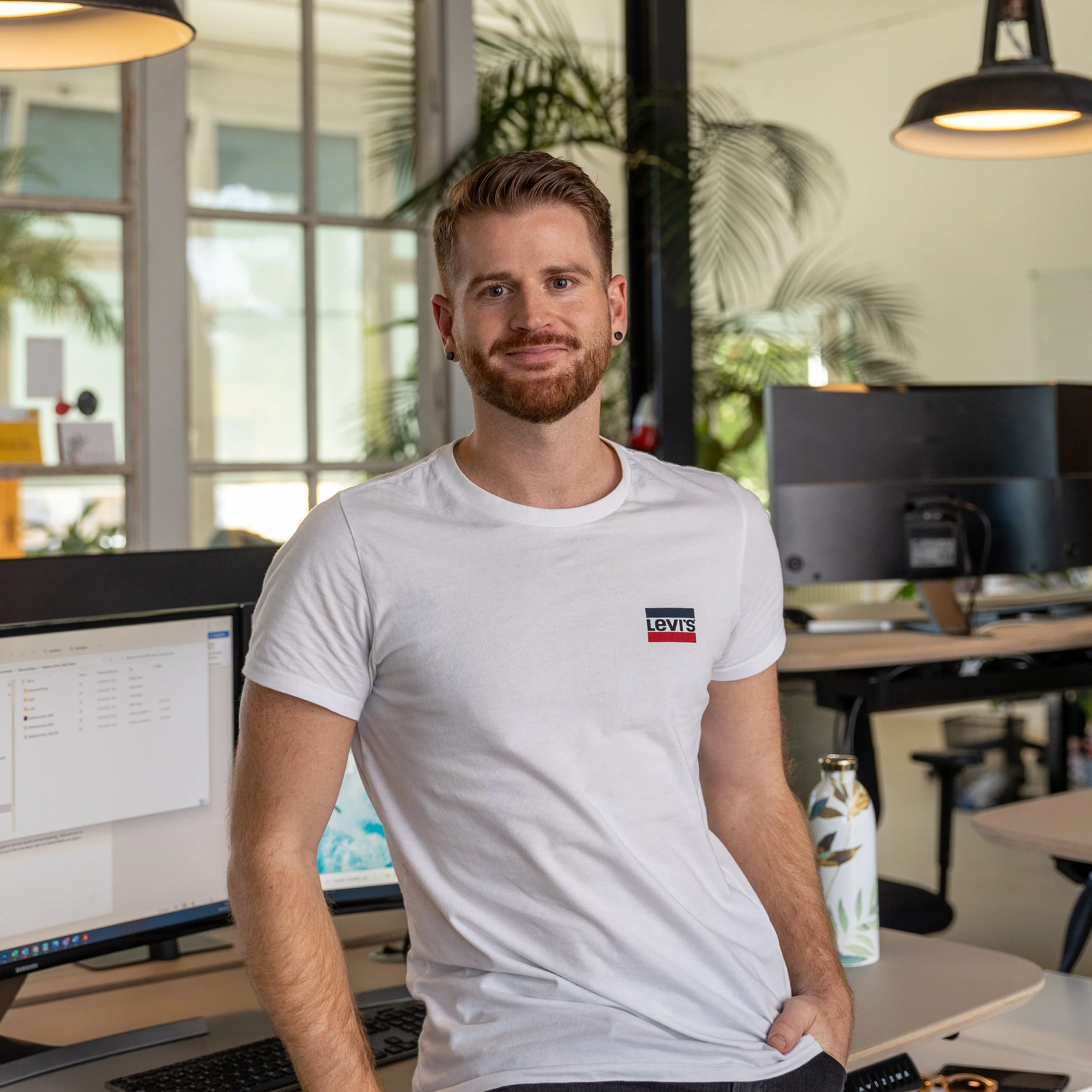 Junger Mann mit roten Haaren und Bart, trägt ein weißes T-Shirt mit Levi's-Logo, steht in einem modernen Büro mit Computern und Pflanzen.