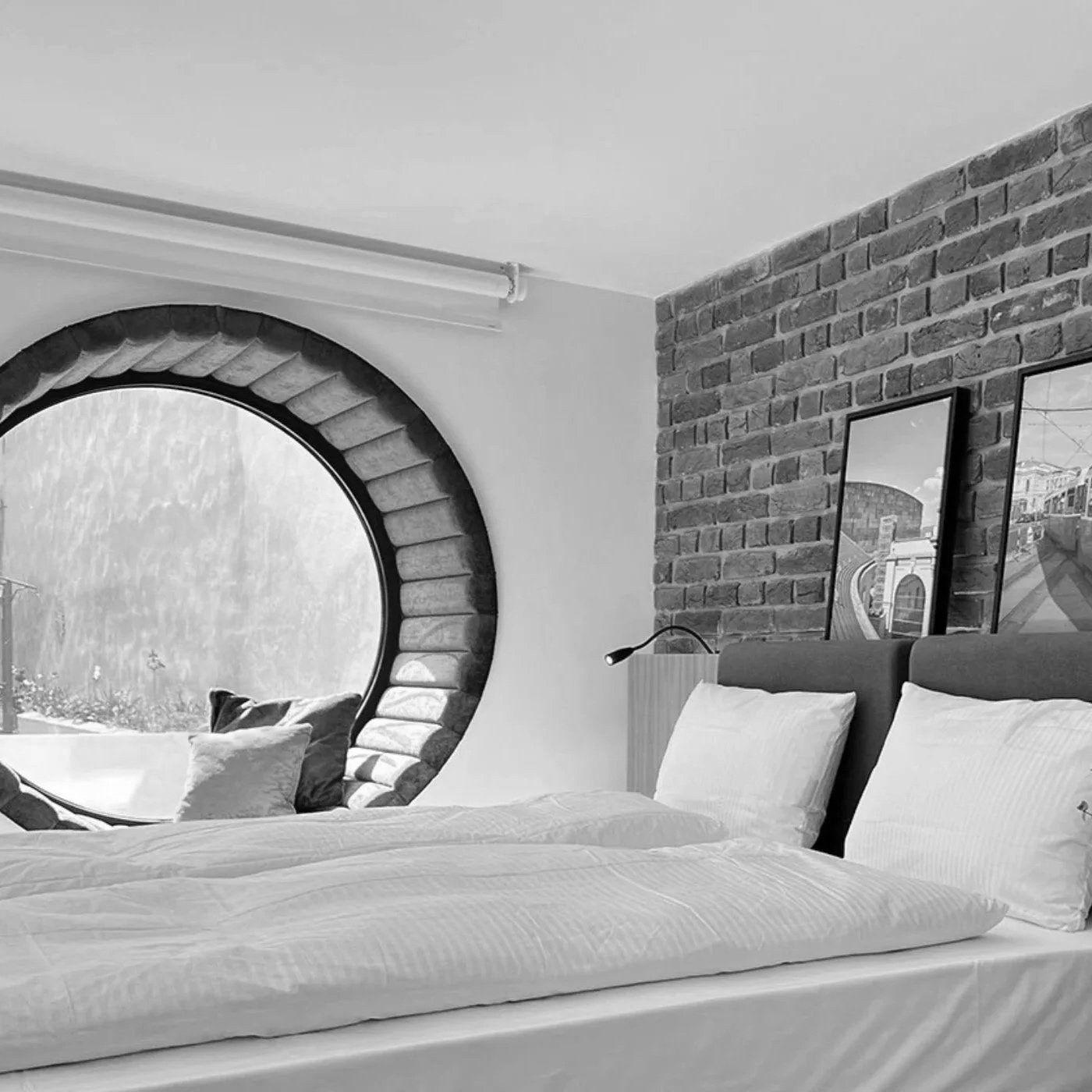 A bedroom with a large round window showing rain outside, brick wall with two framed pictures, bed with white linens and pillows, and a small reading lamp.