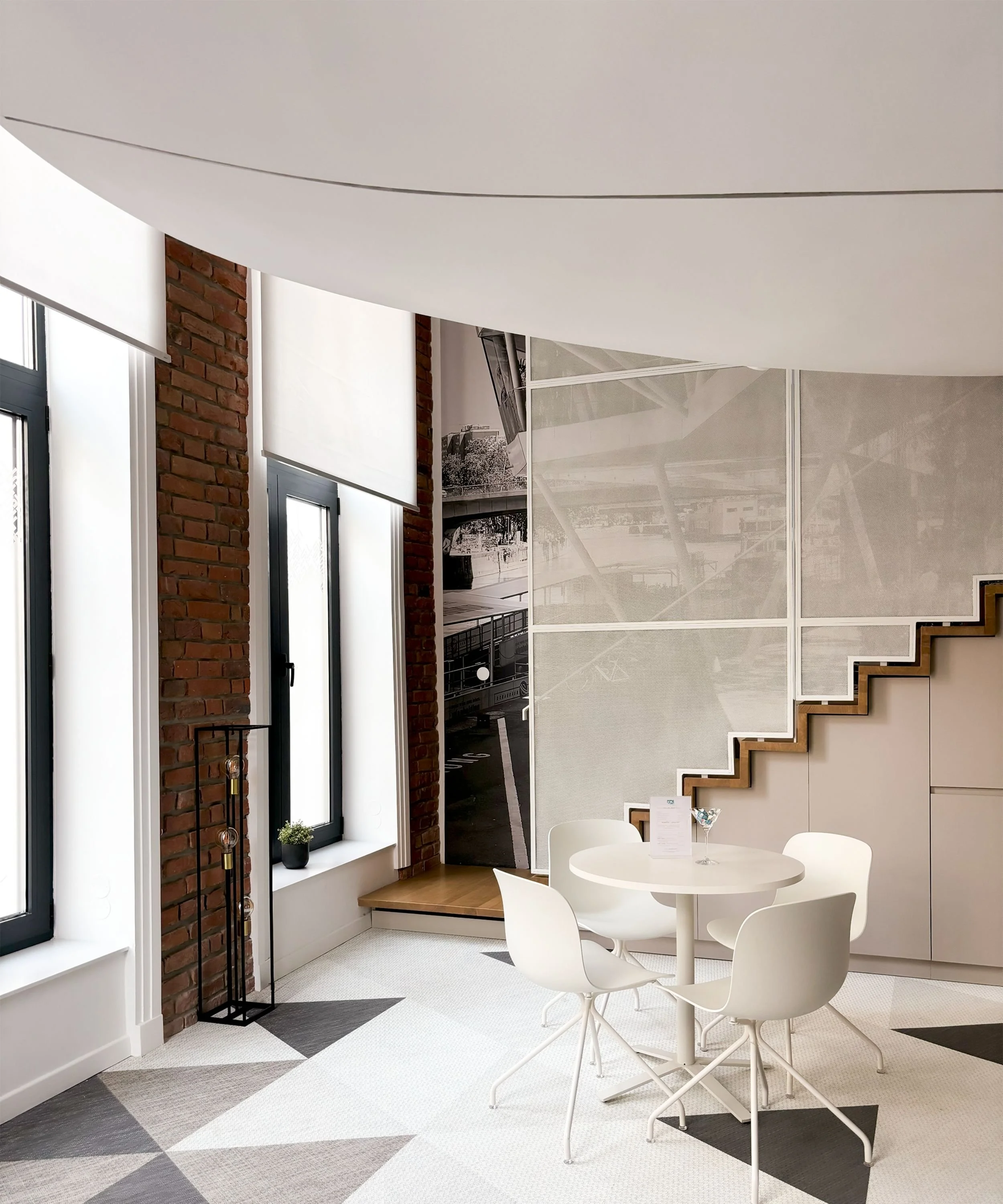 Modern interior with brick columns, large windows with white blinds, a round table with four white chairs, and a staircase with wooden steps.