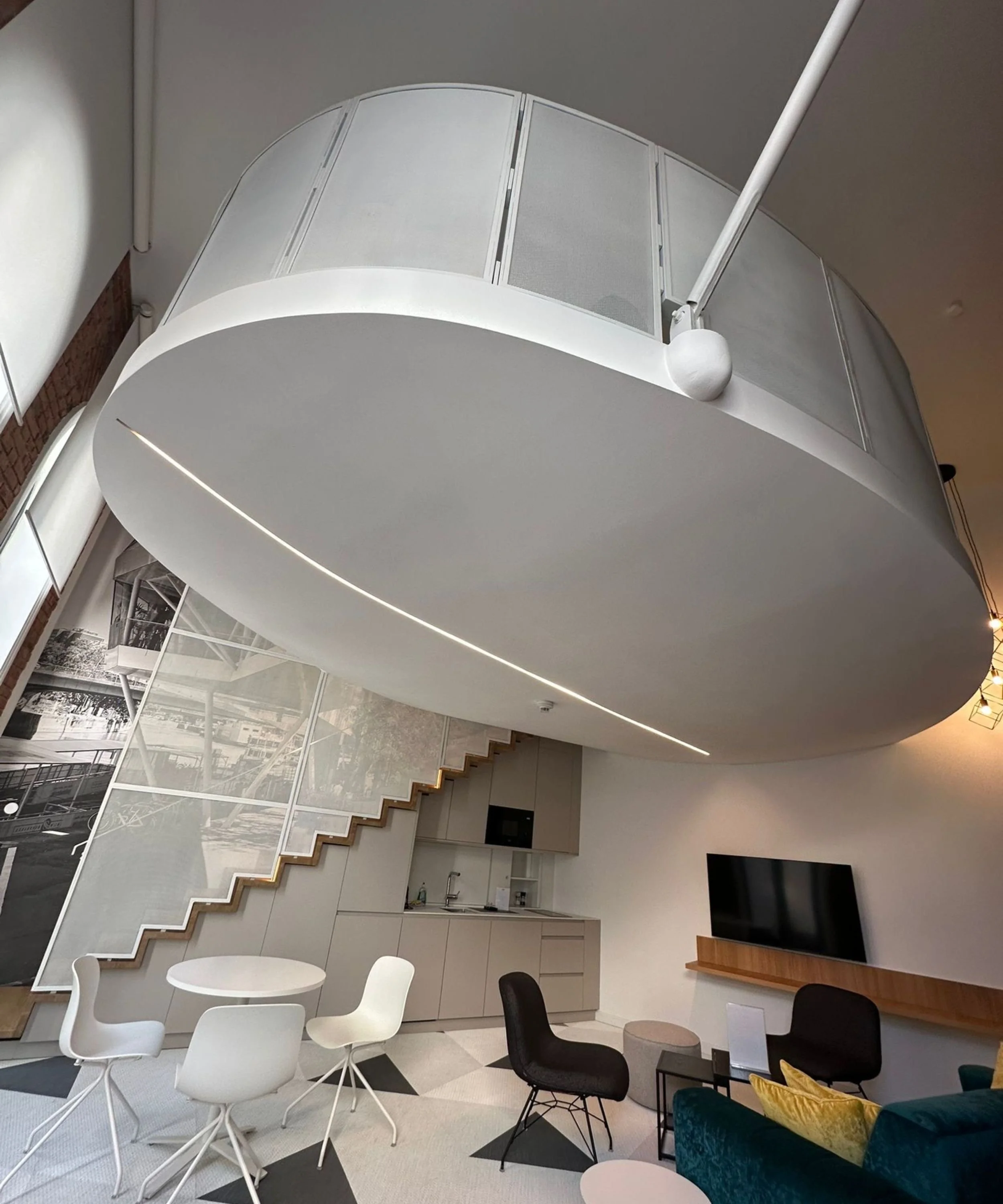 Modern living room with a large white floating ceiling feature, black and white chairs, a TV on a wall-mounted shelf, and a staircase with wooden steps.