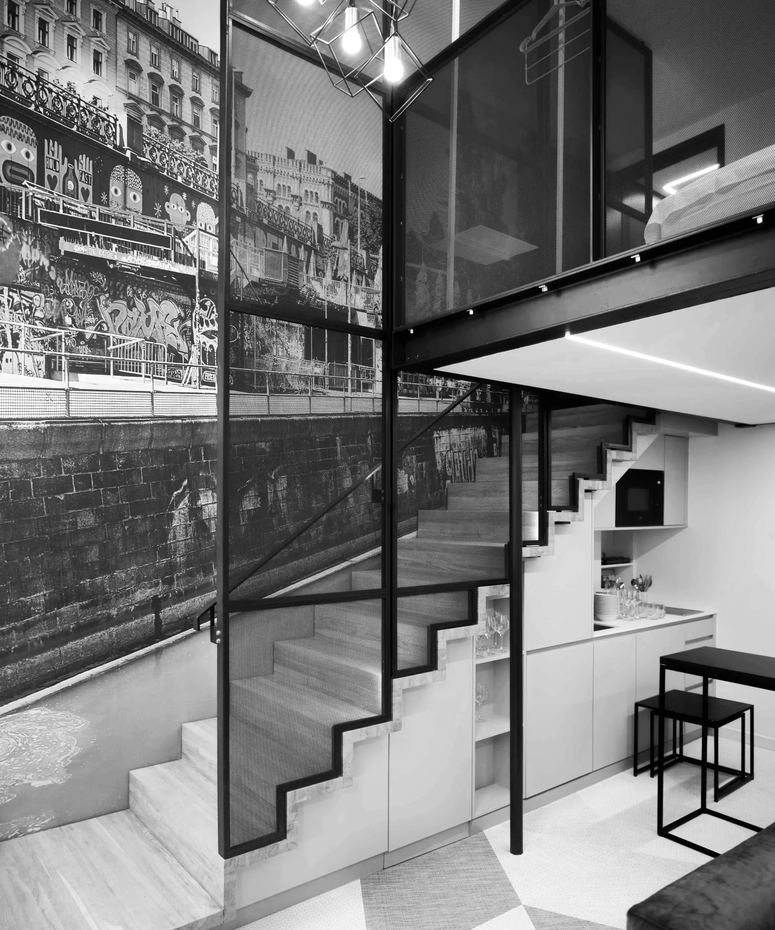 A modern interior with a staircase leading to a lofted area. The wall features a large mural or wallpaper of a cityscape with graffiti, and windows or glass panels above. The space is minimalistic with white cabinetry, glassware, and a small table with stools.