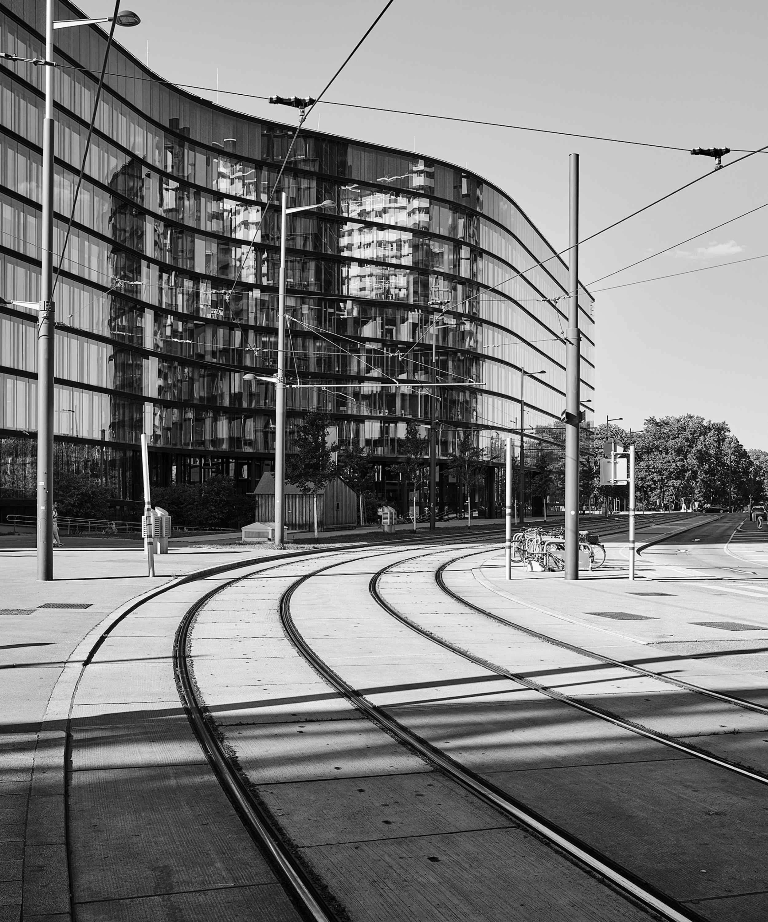 Black and white photo of a modern glass building with curved architecture, tram tracks on the street, and bike racks in the foreground.