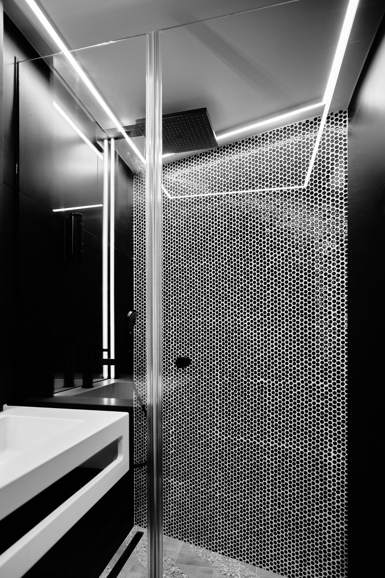 A modern bathroom with black and white decor, featuring a geometric dotted wall pattern, a corner shower with glass and metal accents, a white sink, and LED strip lighting on the ceiling.