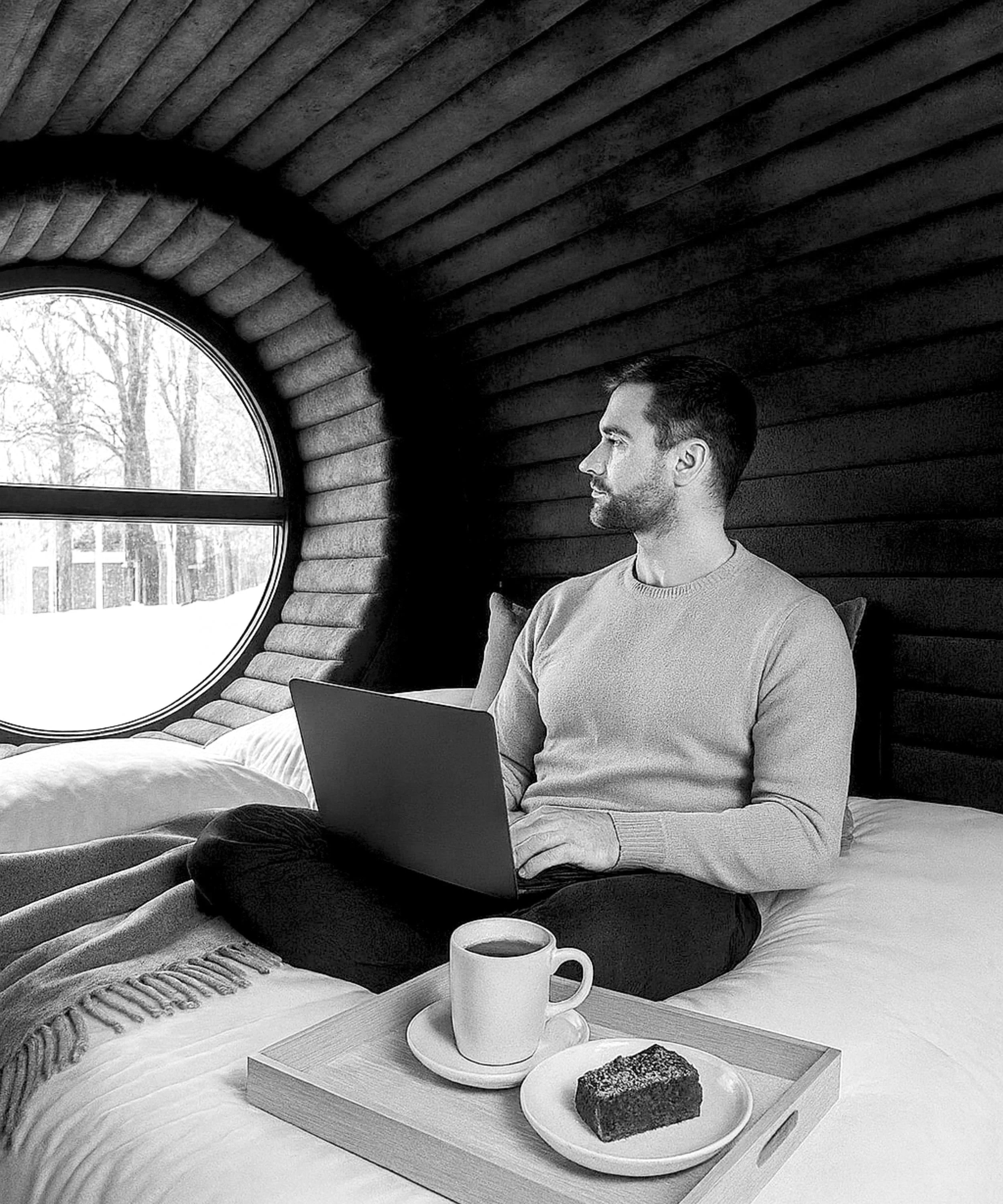 A man sitting cross-legged on a bed with a laptop on his lap, a tray with a cup of coffee and a piece of cake in front of him, inside a room with wooden panel walls and a round window showing a wintery outdoor scene.