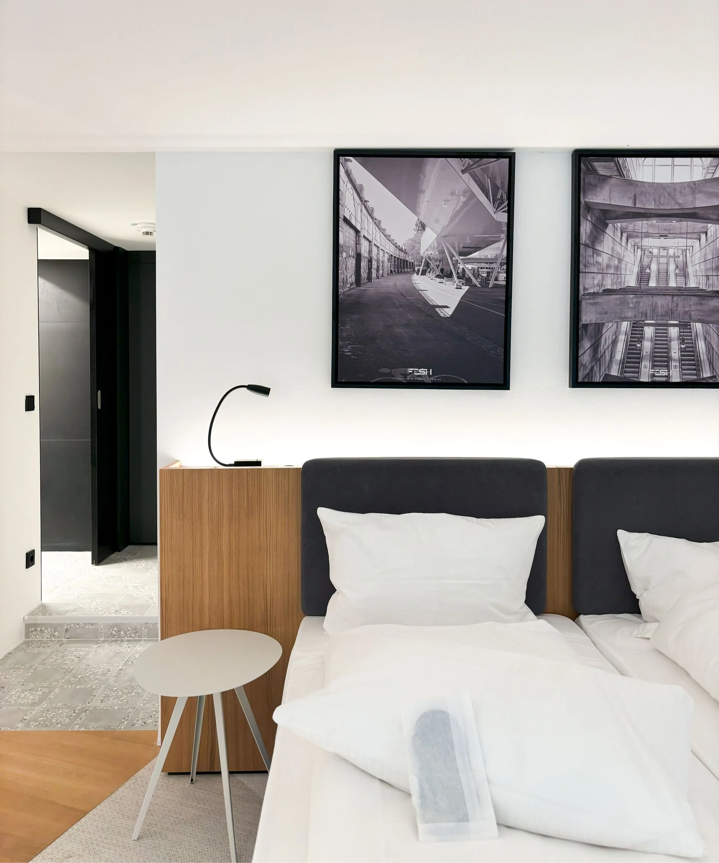 A modern hotel room with a bed, white bedding and pillows, a round side table, a black reading lamp on a wooden headboard, and framed black-and-white architectural photographs on the wall.