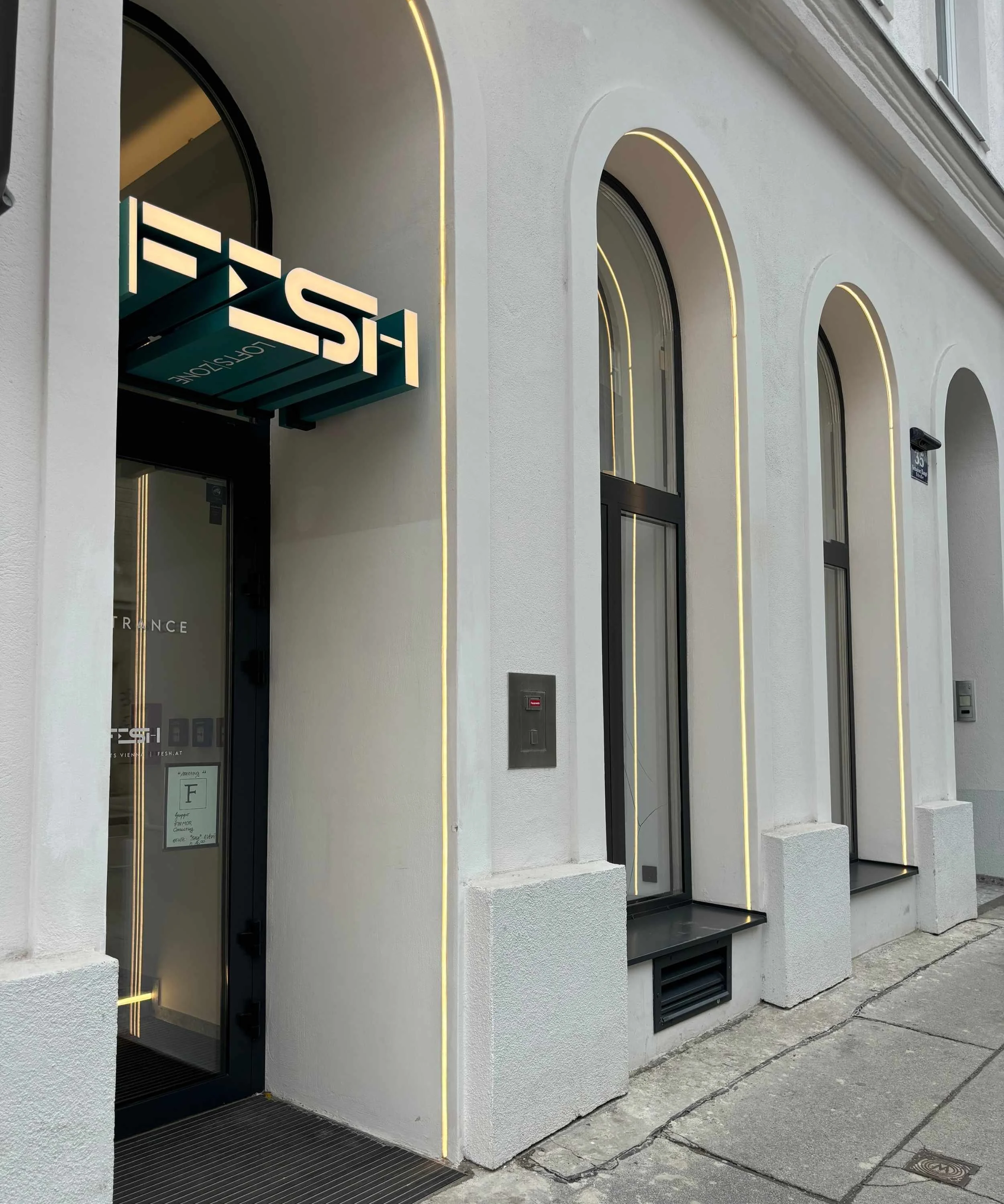 Modern building exterior with arched windows outlined with yellow neon lights and a black door with glass that has the word 'ENTRANCE' on it. A neon sign above the door reads 'FESH' or similar.