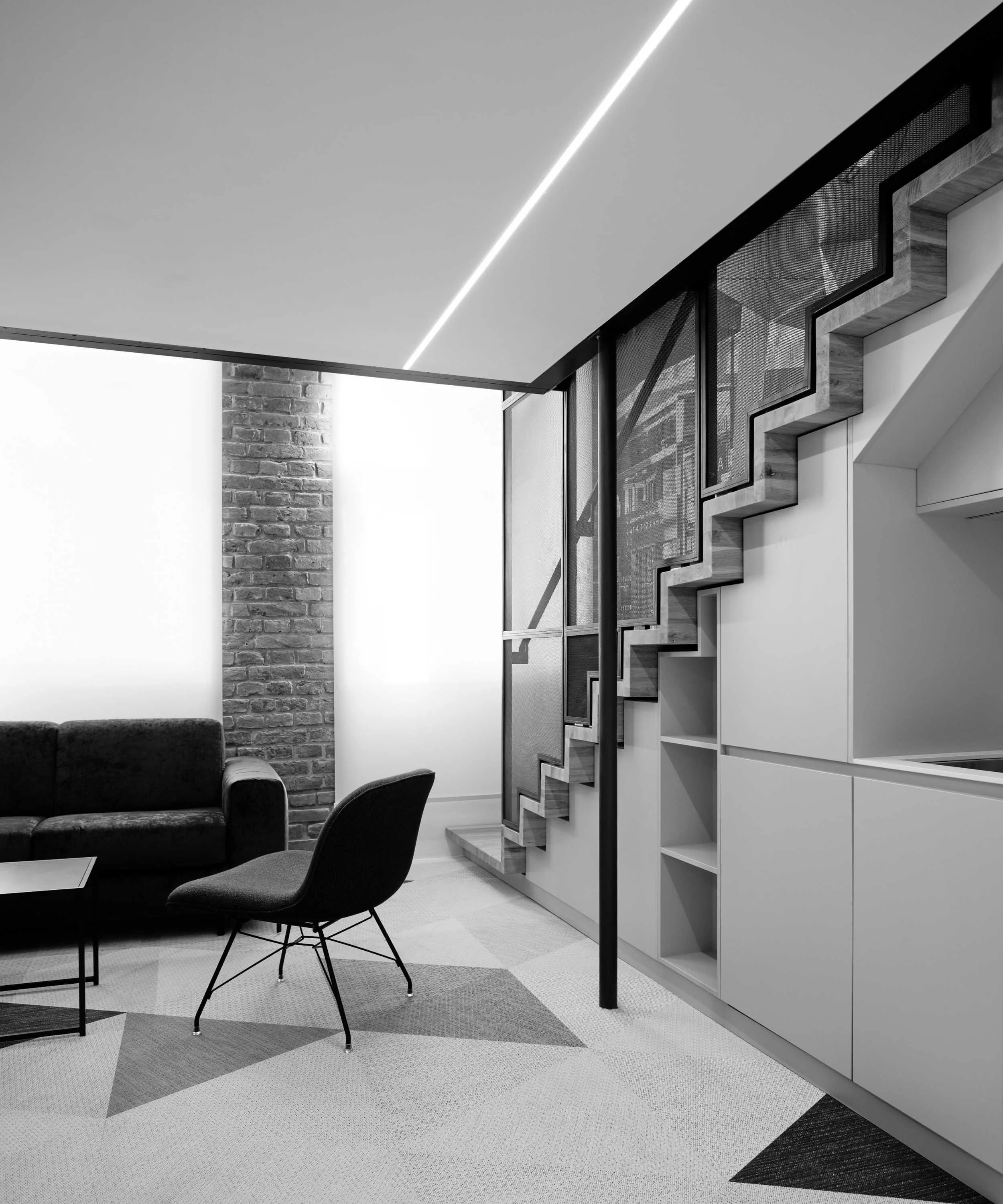 Modern living space with a black sofa, a chair, a coffee table, and a staircase with a unique zigzag shadow pattern, illuminated by a sleek ceiling light, with a brick accent wall.