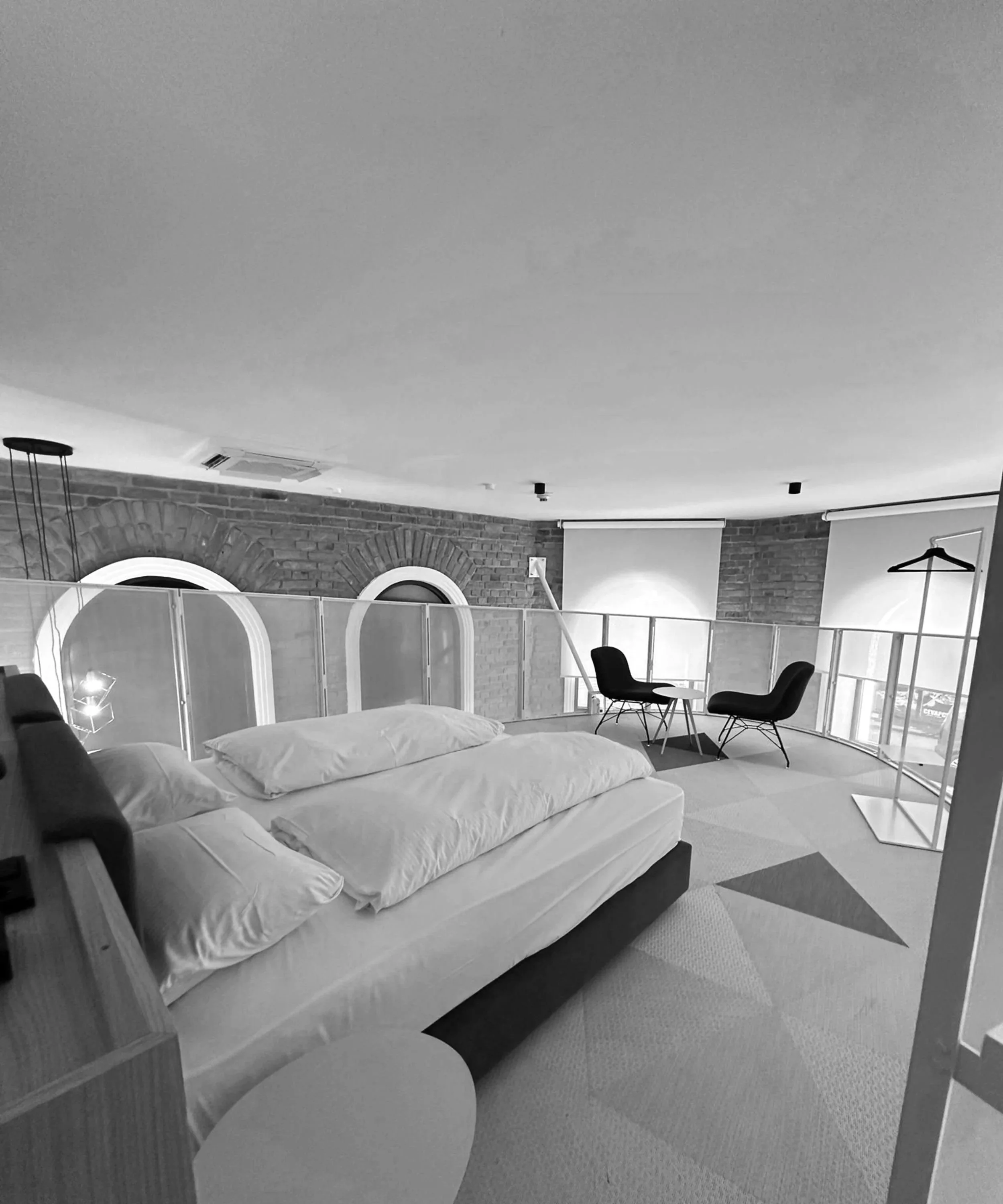 A black and white photo of a cozy bedroom with a bed, two chairs, and a small round table on a rug near a large window with a brick wall background.