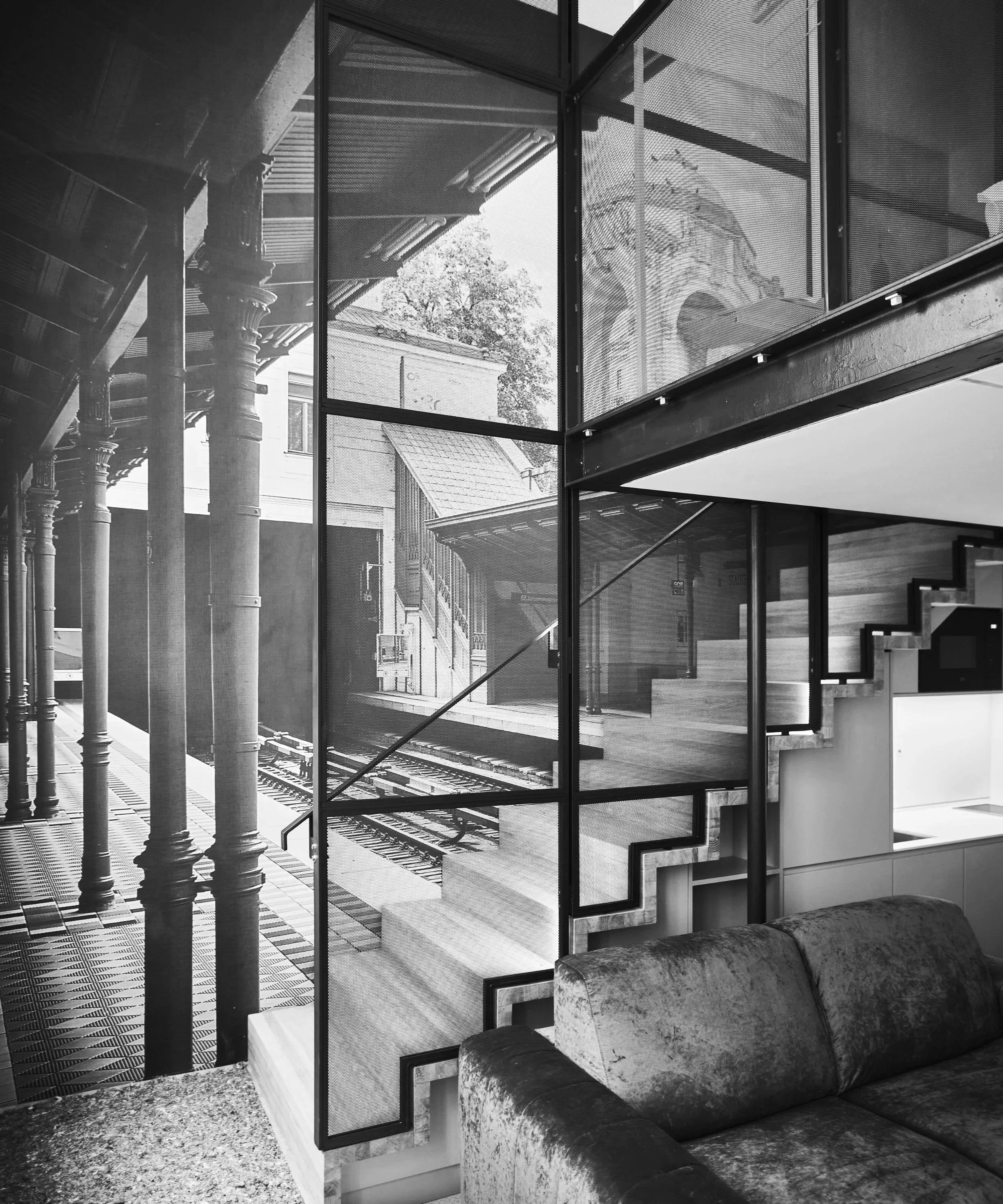 Interior of a modern building with glass walls, stairs, and a velvet sofa, showing an external train station platform through the glass.