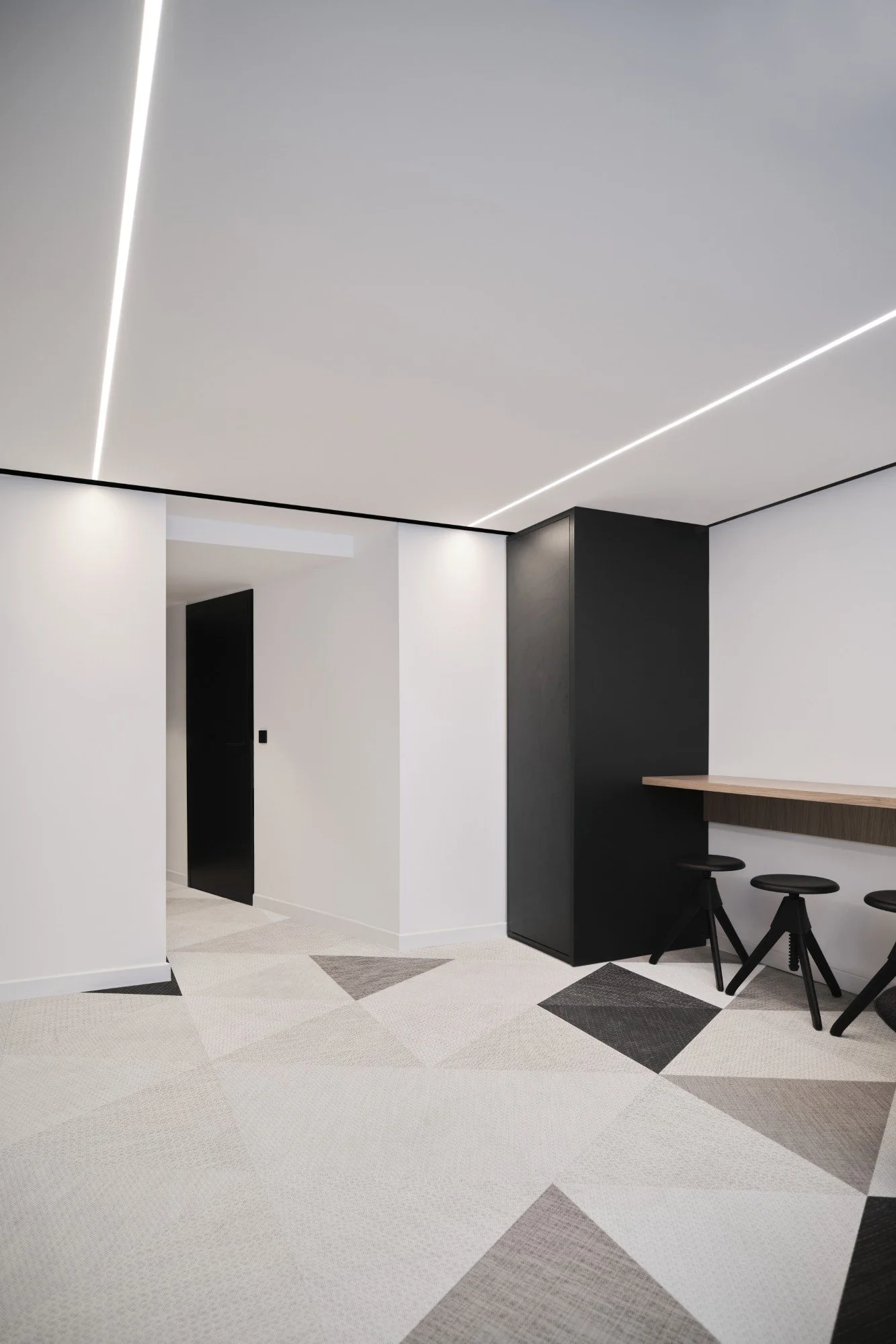 Modern minimalist interior with white walls, a black cabinet, a wooden counter with black stools, and geometric patterned carpet.