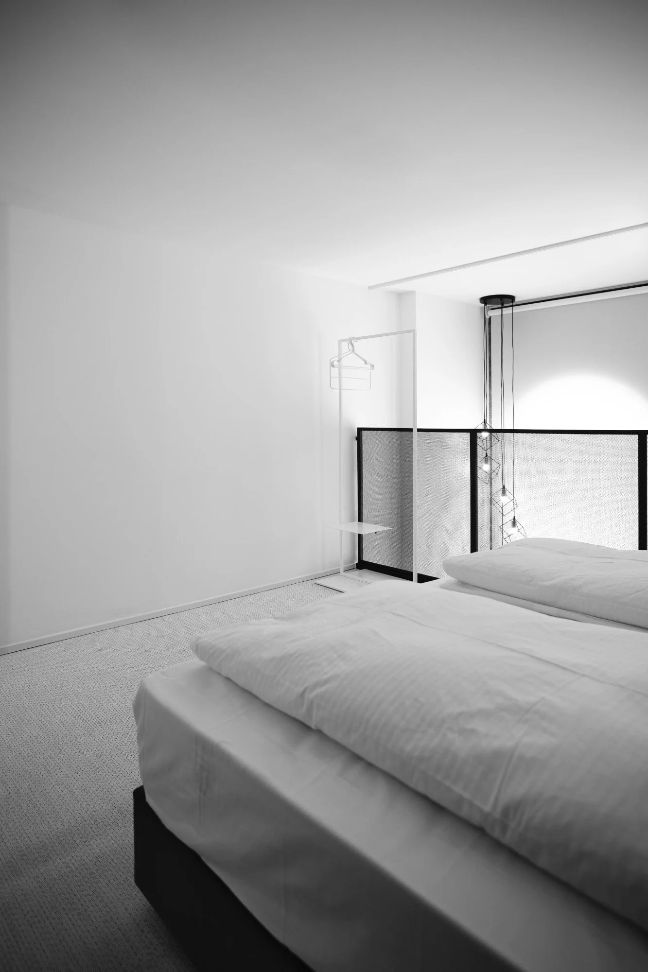 A minimalist bedroom with two neatly made beds, white bedding, and a black headboard, featuring modern pendant lights and a small clothing rack near the window.