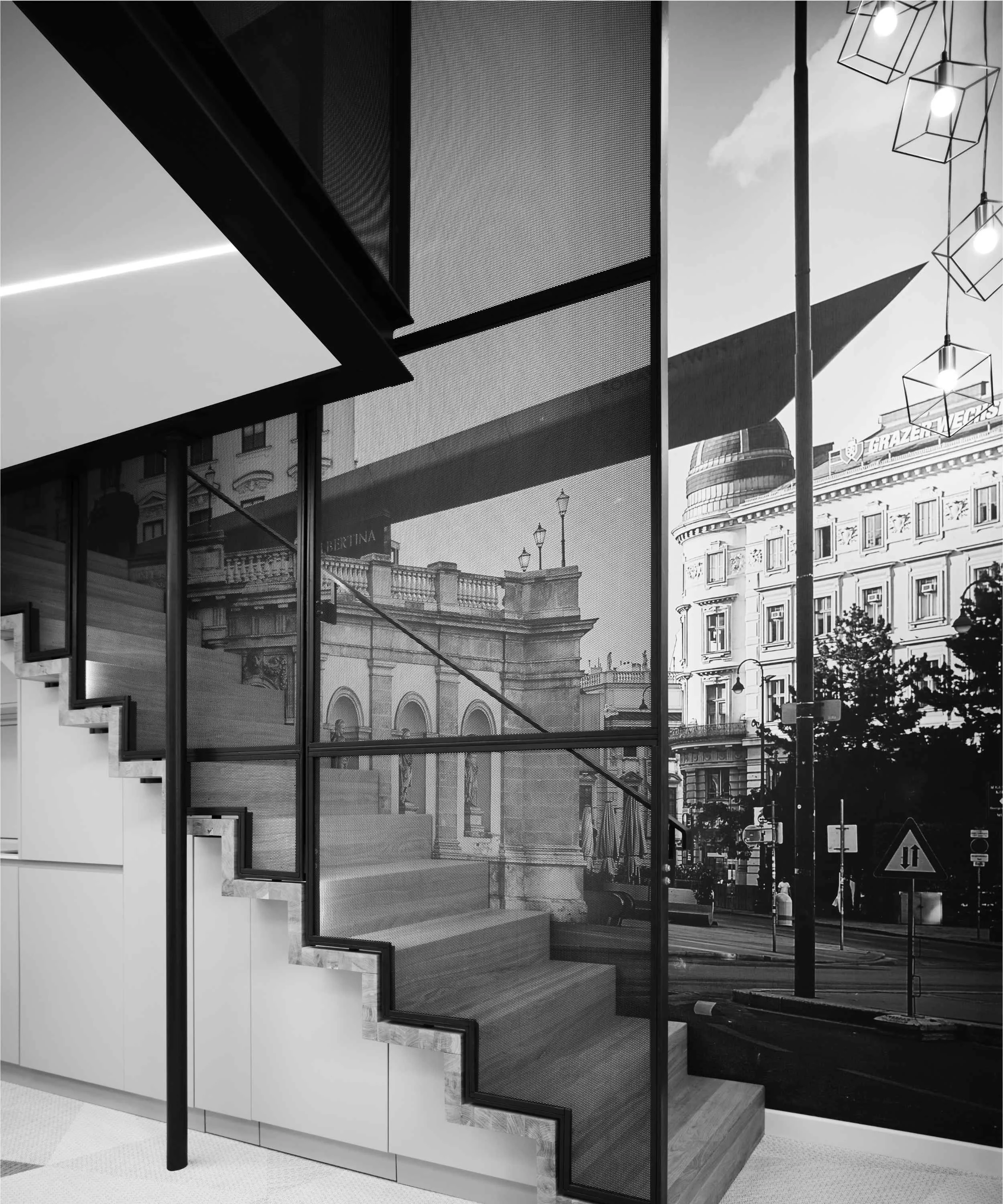 Black and white photo of a modern interior staircase with glass walls, overlooking a city street scene with historic buildings and street lamps.