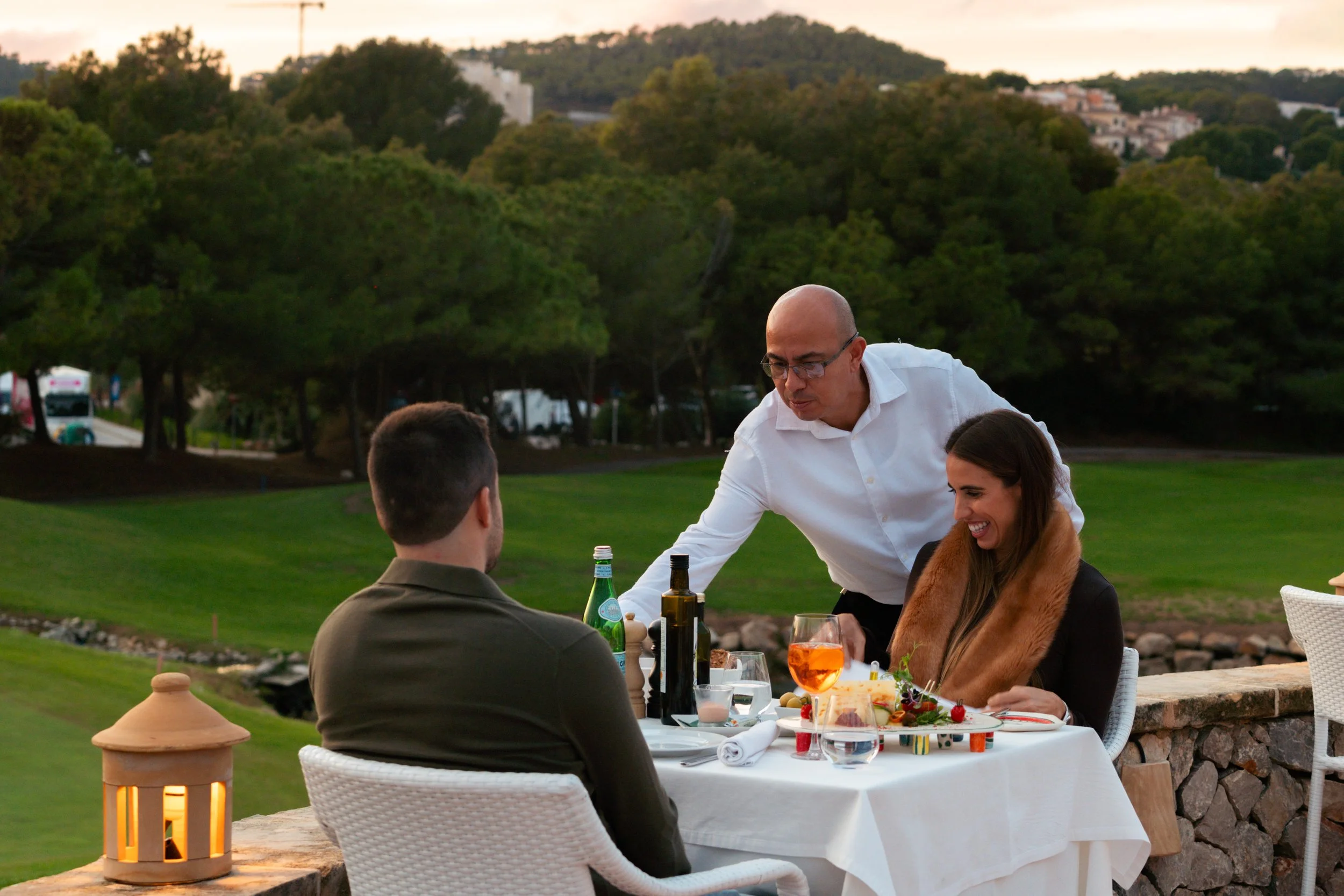 Tres personas en una cena al aire libre en un campo con árboles y colinas: un hombre sirviendo comida a una mujer que sonríe, mientras un hombre con cabello corto observa. La mesa tiene comida, bebidas y una linterna de vela.