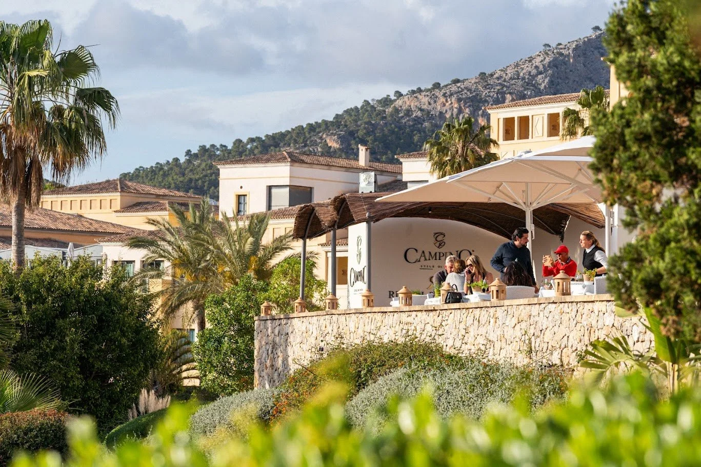 Clientes disfrutando en una terraza al aire libre en un restaurante con vista a montañas, rodeada de vegetación y palmeras.
