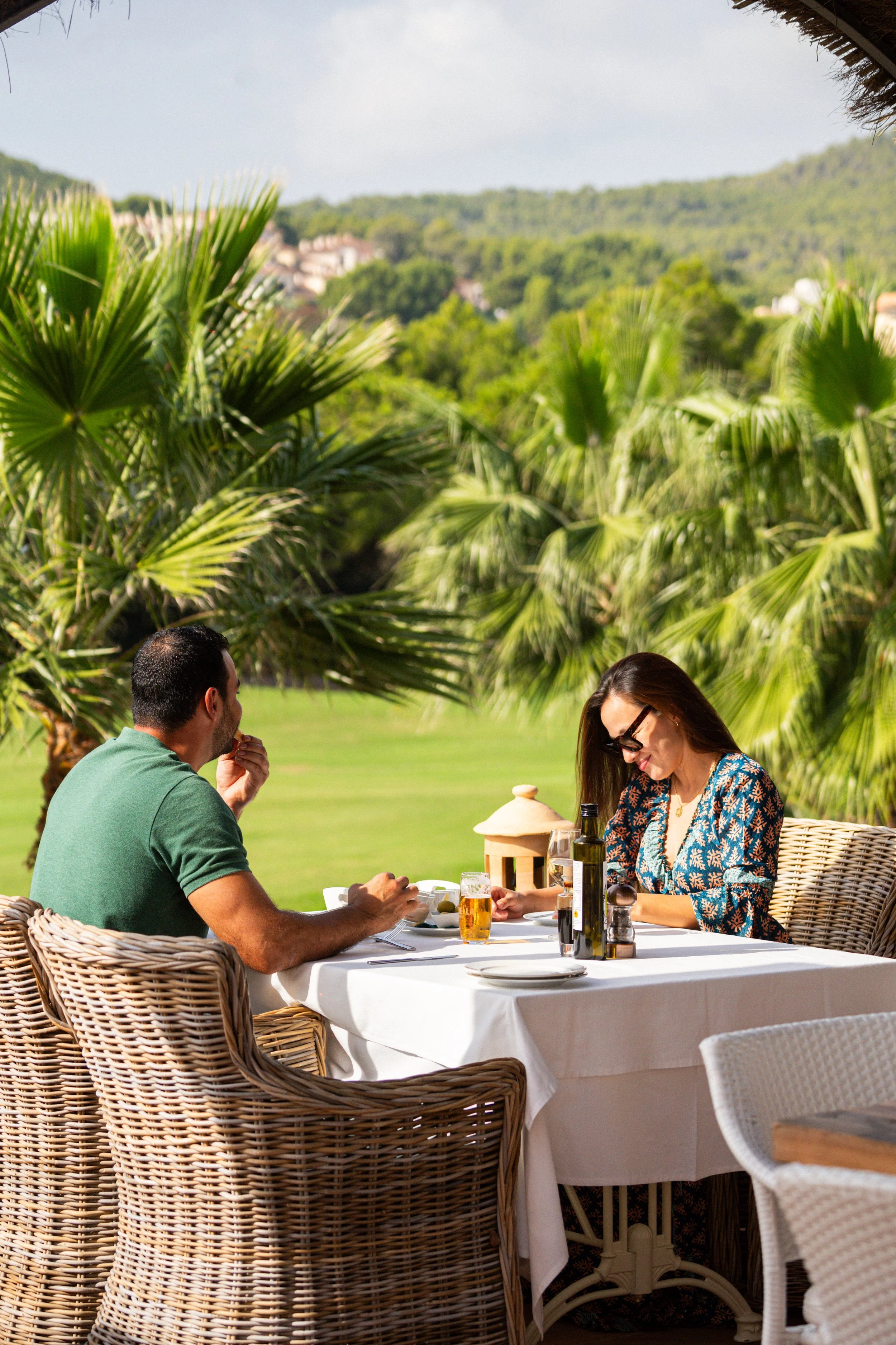 Dos personas cenando en un restaurante al aire libre con fondo de vegetación tropical y colinas.