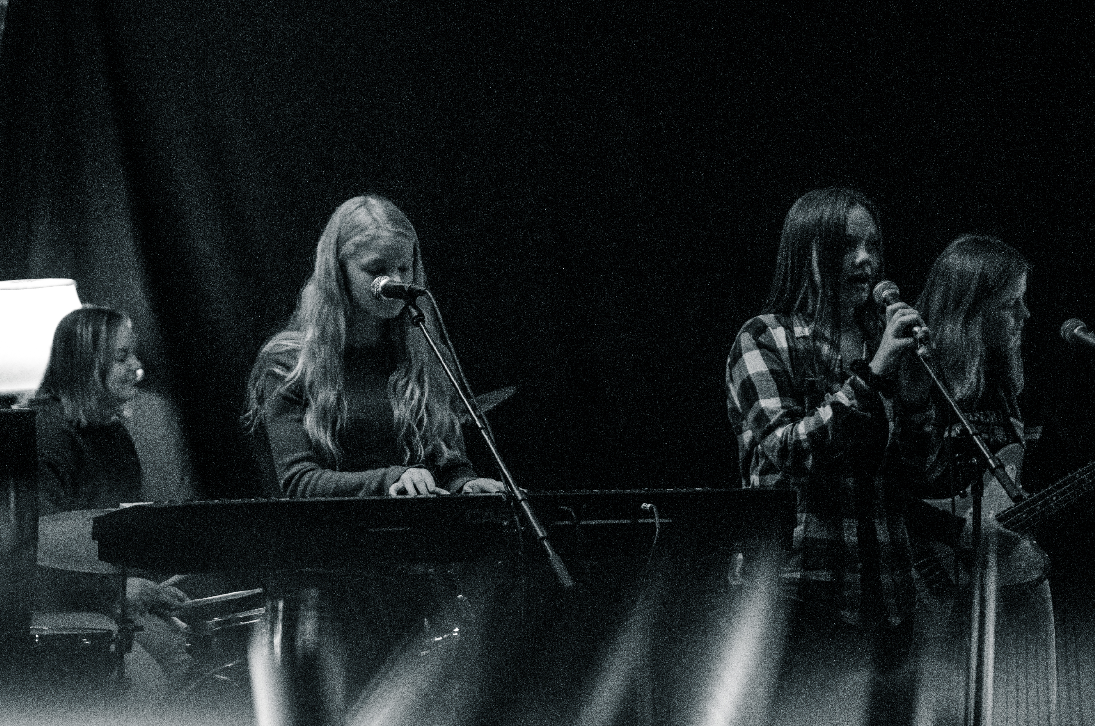 Et mørkt, fargeløst bilde av fire musikere på en scene. En spiller synth, en synger, en spiller gitar og en spiller trommer.