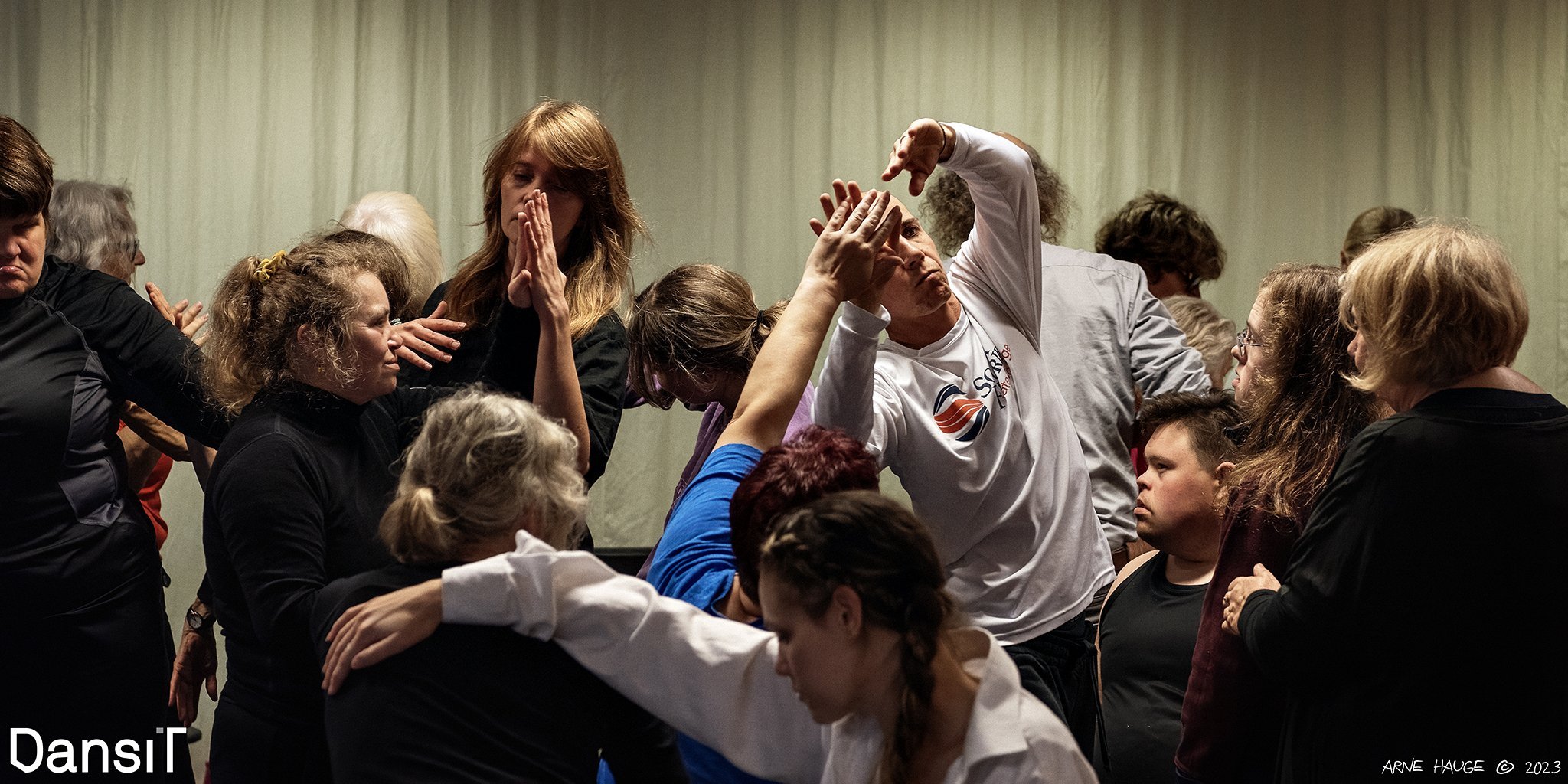 I underkant av 20 mennesker danser ekspressivt i et rom. To par holder håndflatene mot hverandre, mens de andre danserne er vendt i ulike retninger av hverandre. Ingen ser i kamera.