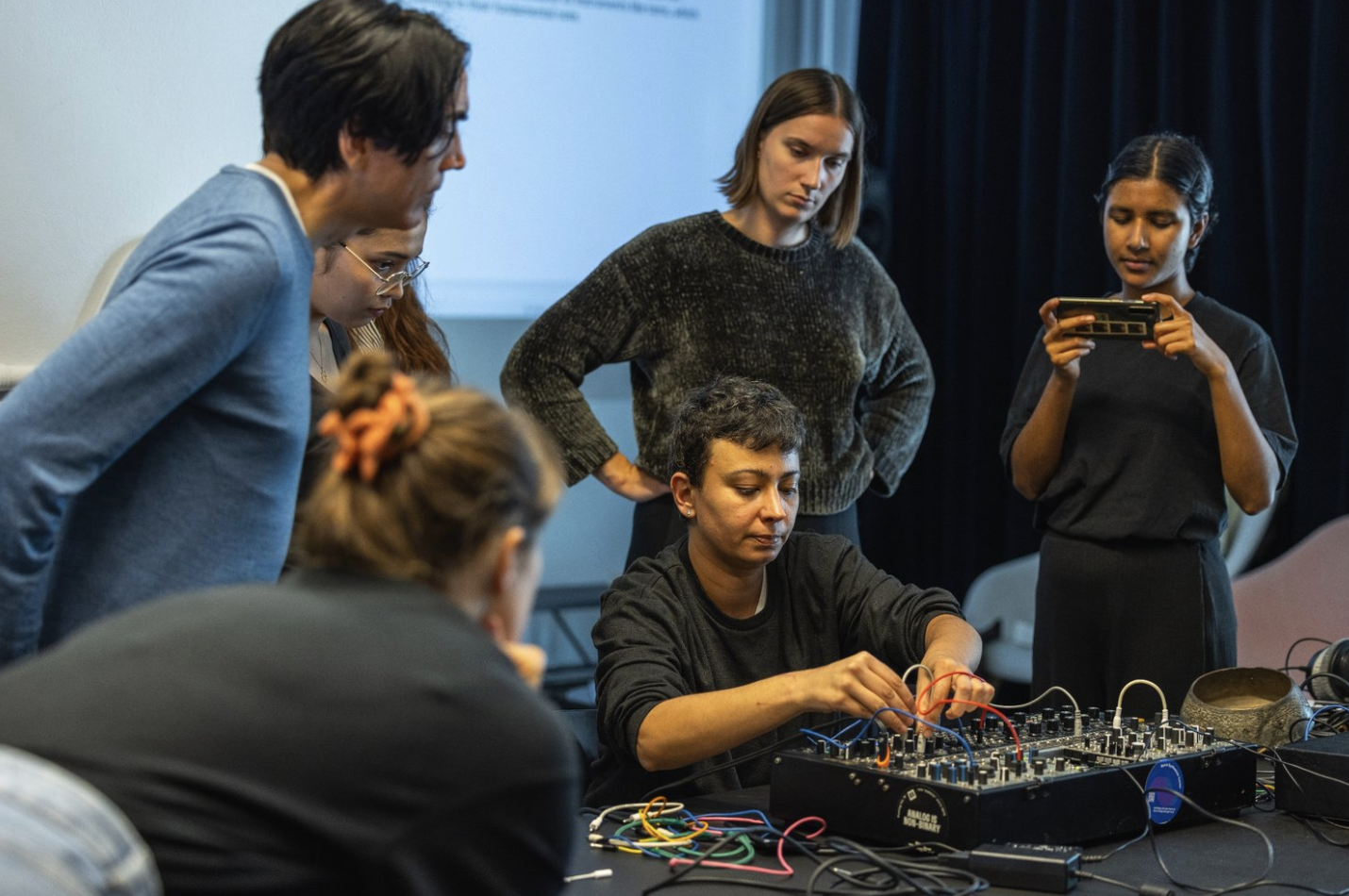 Fem personer samlet rundt et bord der en justerer kabler på en elektronisk lydmikser, mens de andre følger med.