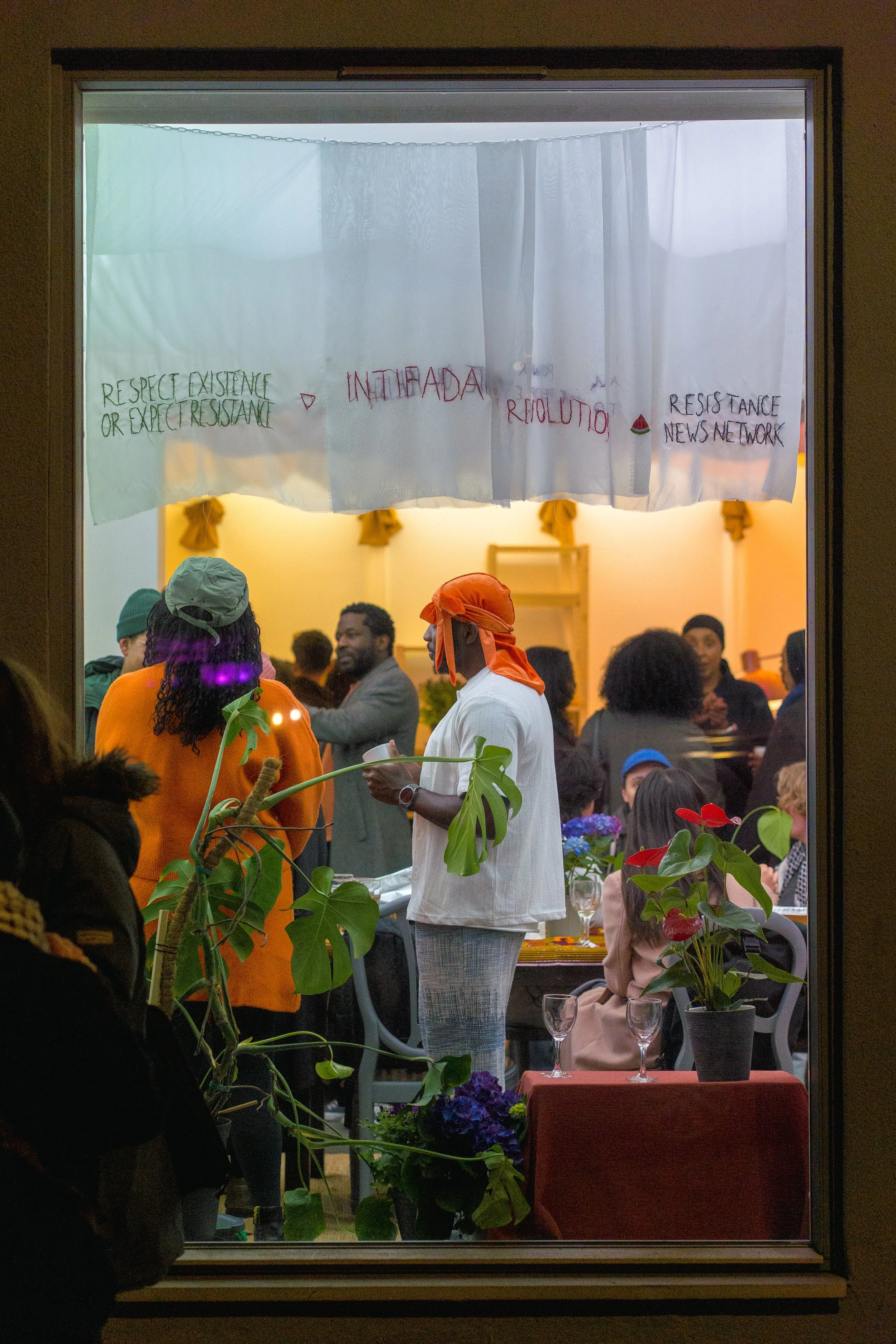 Et innblikk gjennom et vindu viser mennesker samlet i et lyst lokale, omgitt av planter og broderte gardiner.