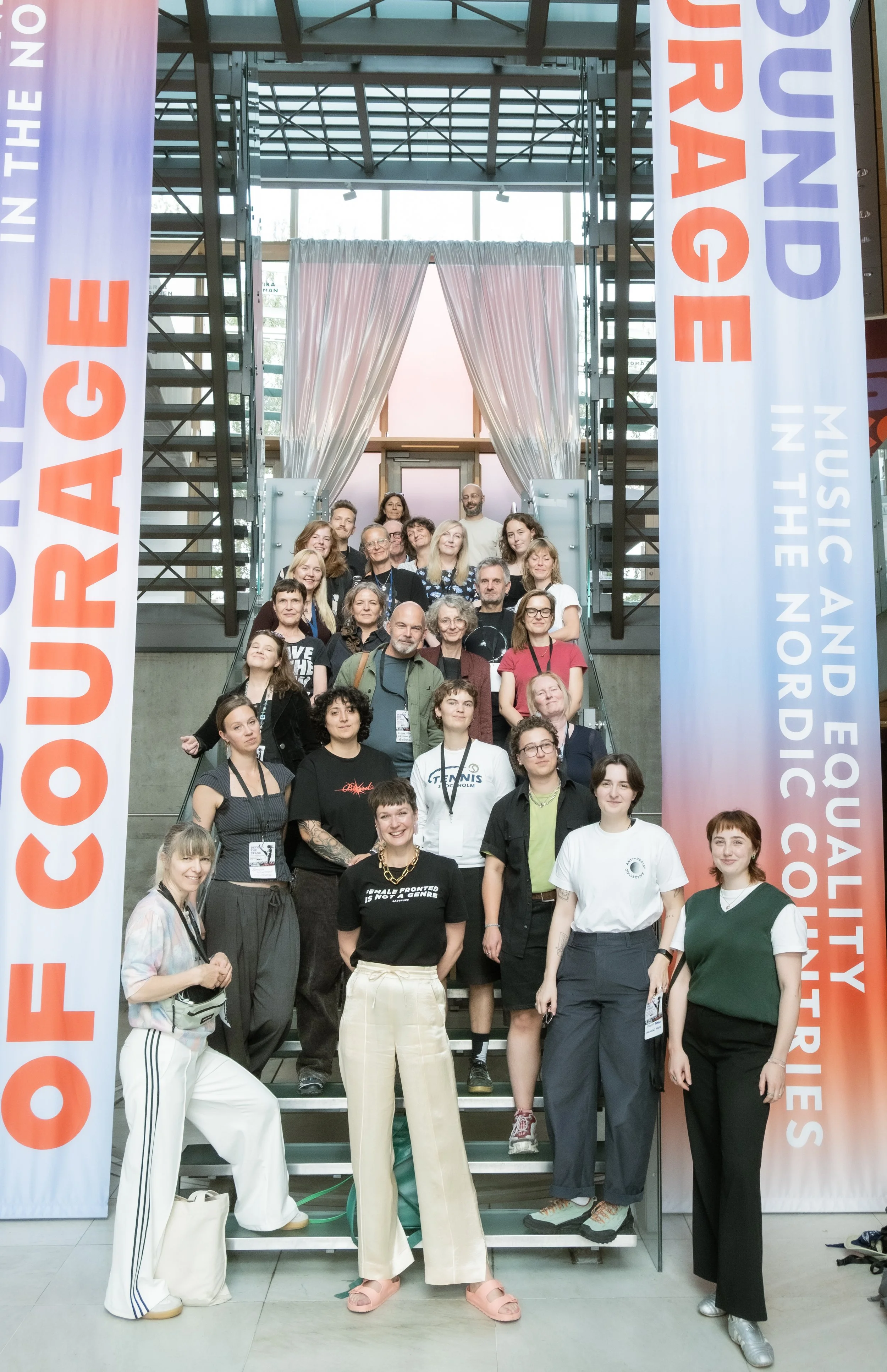 Gruppefoto av deltakere på «Beyond the Stage» i Felleshus i Berlin. Rundt 20 personer står samlet i en trapp mellom to store bannere for utstillingen «The Sound of Courage – Music and Equality in the Nordic Countries».