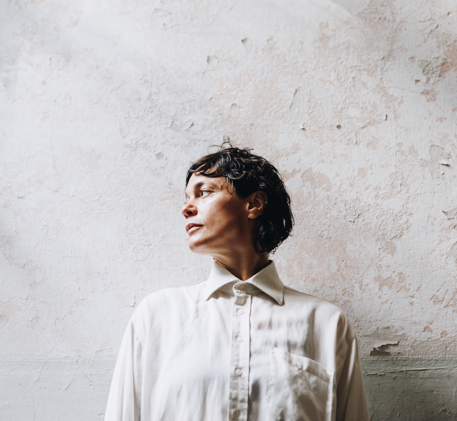 Mia Habib står i hvit skjorte foran en lys rustikk vegg.  Hun ser til siden og har et ettertenksomt uttrykk.