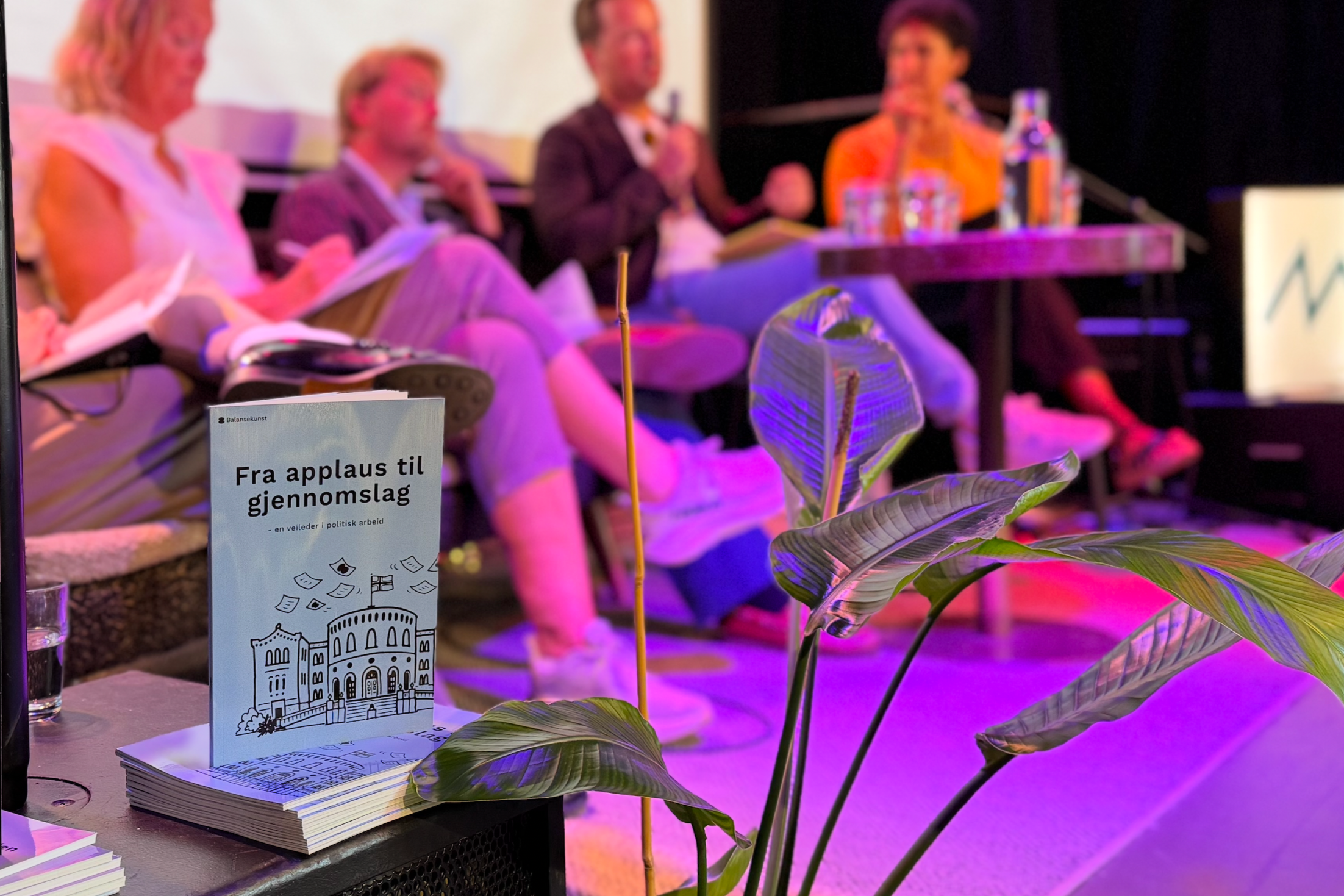 Panelsamtale på en scene, med brosjyren «Fra applaus til gjennomslag» og en grønn plante i forgrunnen.