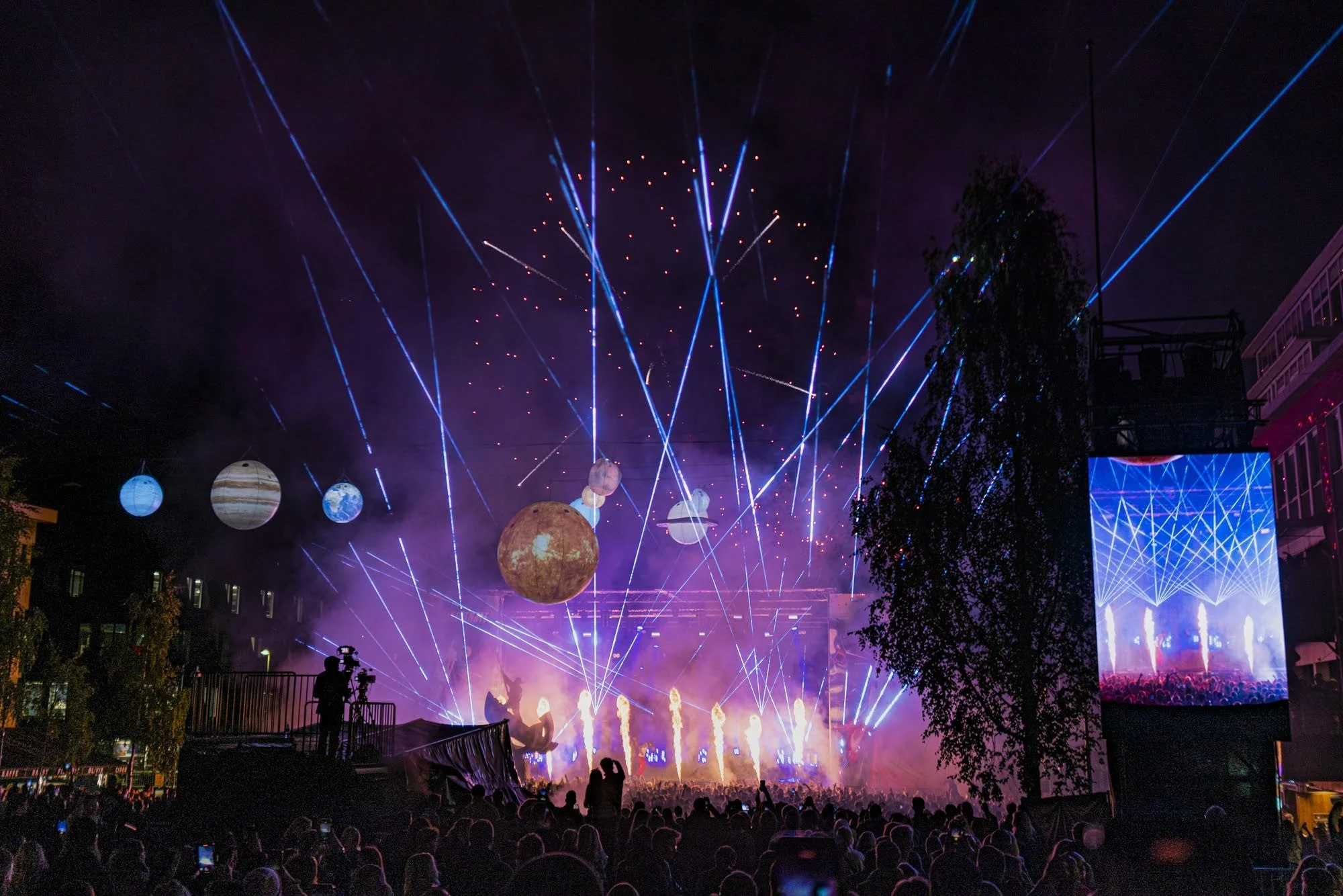 Konsertfoto som viser et forkehav foran en stor scene med masse lyseffekter, pyroshow og rekvisitter som ser ut som planeter.