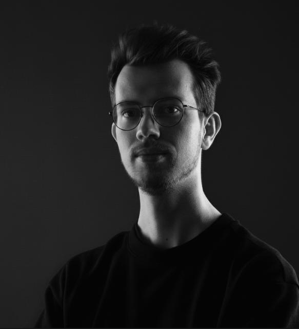 Portretfoto van een jonge man met bruin haar, bril en een zwart shirt, in zwart-wit, op een donkere achtergrond.