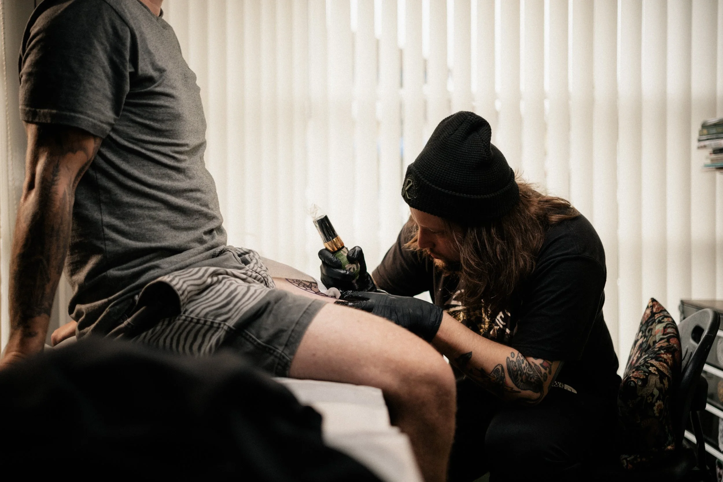 A tattoo artist wearing a black beanie and gloves is tattooing a man's thigh in a room with vertical blinds. The man is sitting on a bed, wearing a gray t-shirt and striped shorts.