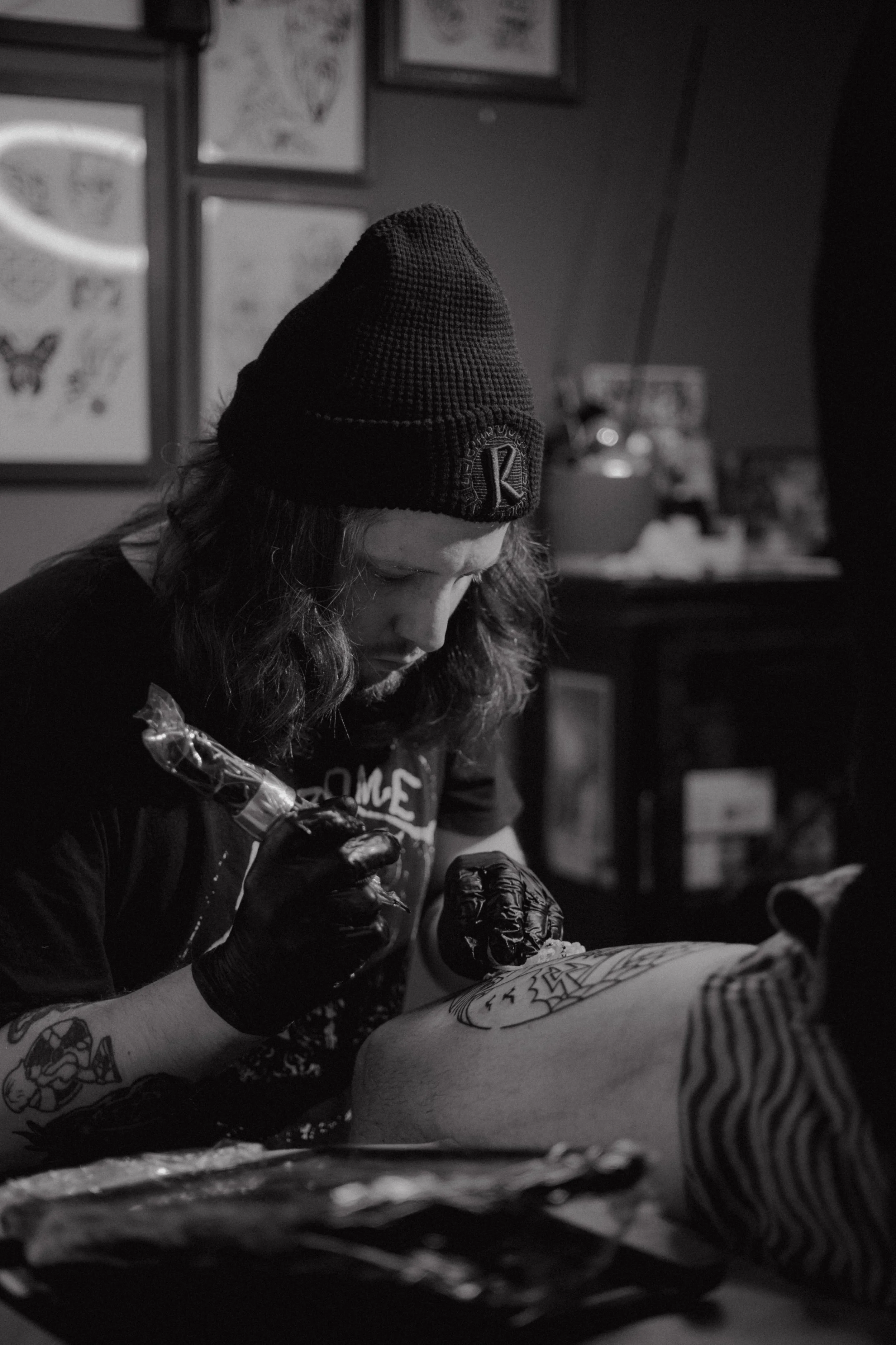 A person with long hair, wearing a beanie and black gloves, is tattooing on another person's back in a tattoo studio.