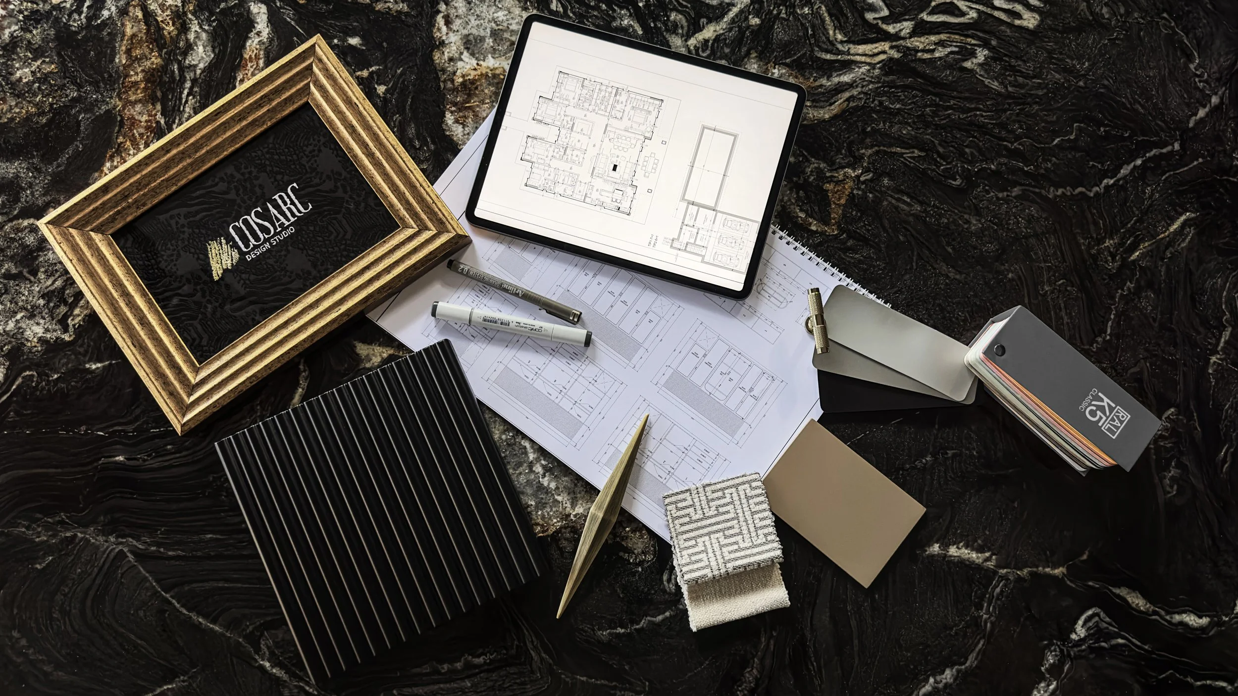 Design studio workspace with architectural plans, color swatches, fabric samples, a tablet displaying a building layout, and various design tools on a dark, marble surface.