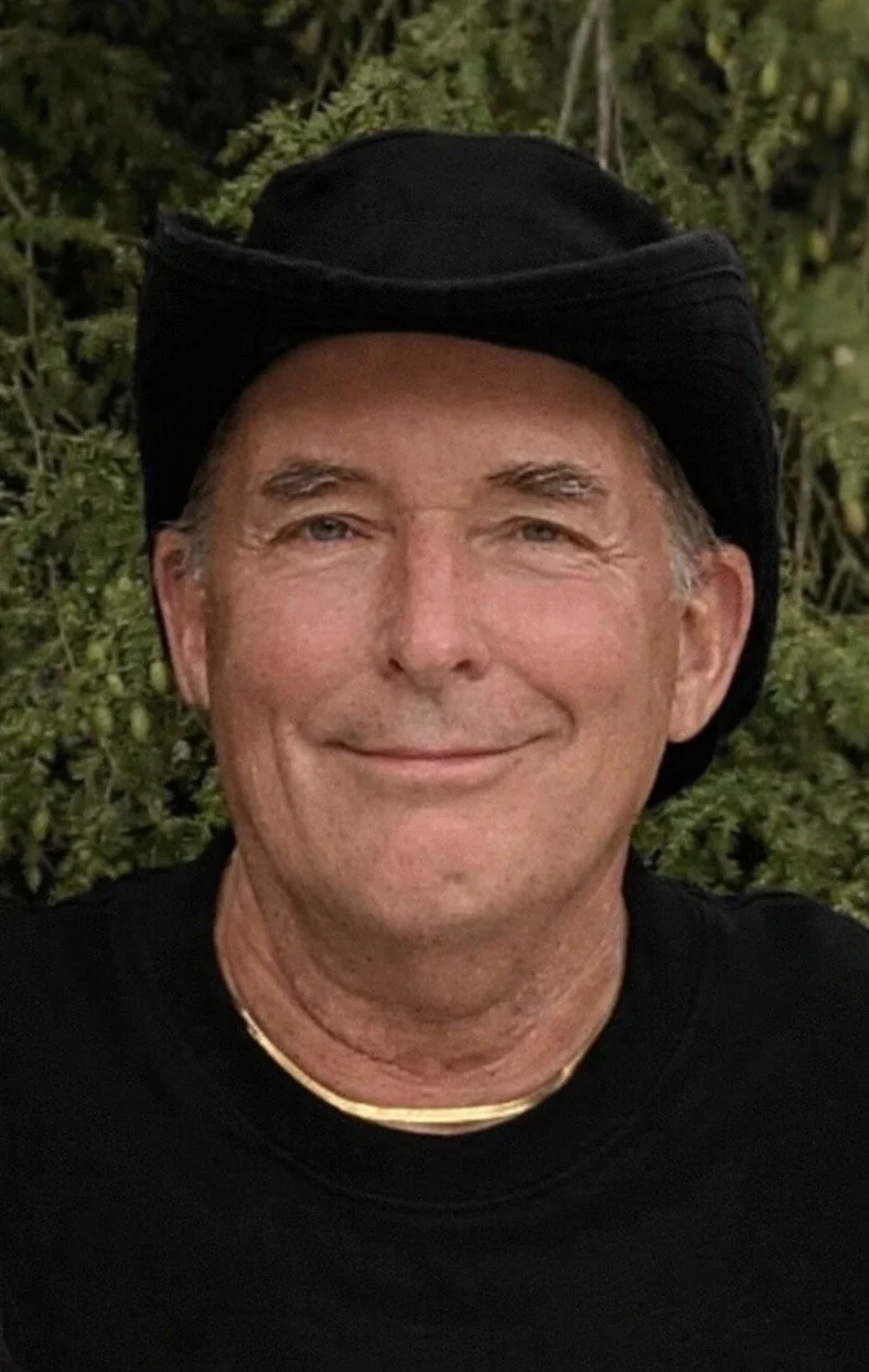 Man smiling, wearing a cowboy hat with an American flag pattern and a navy blue shirt with white stars, standing outdoors with greenery in the background.