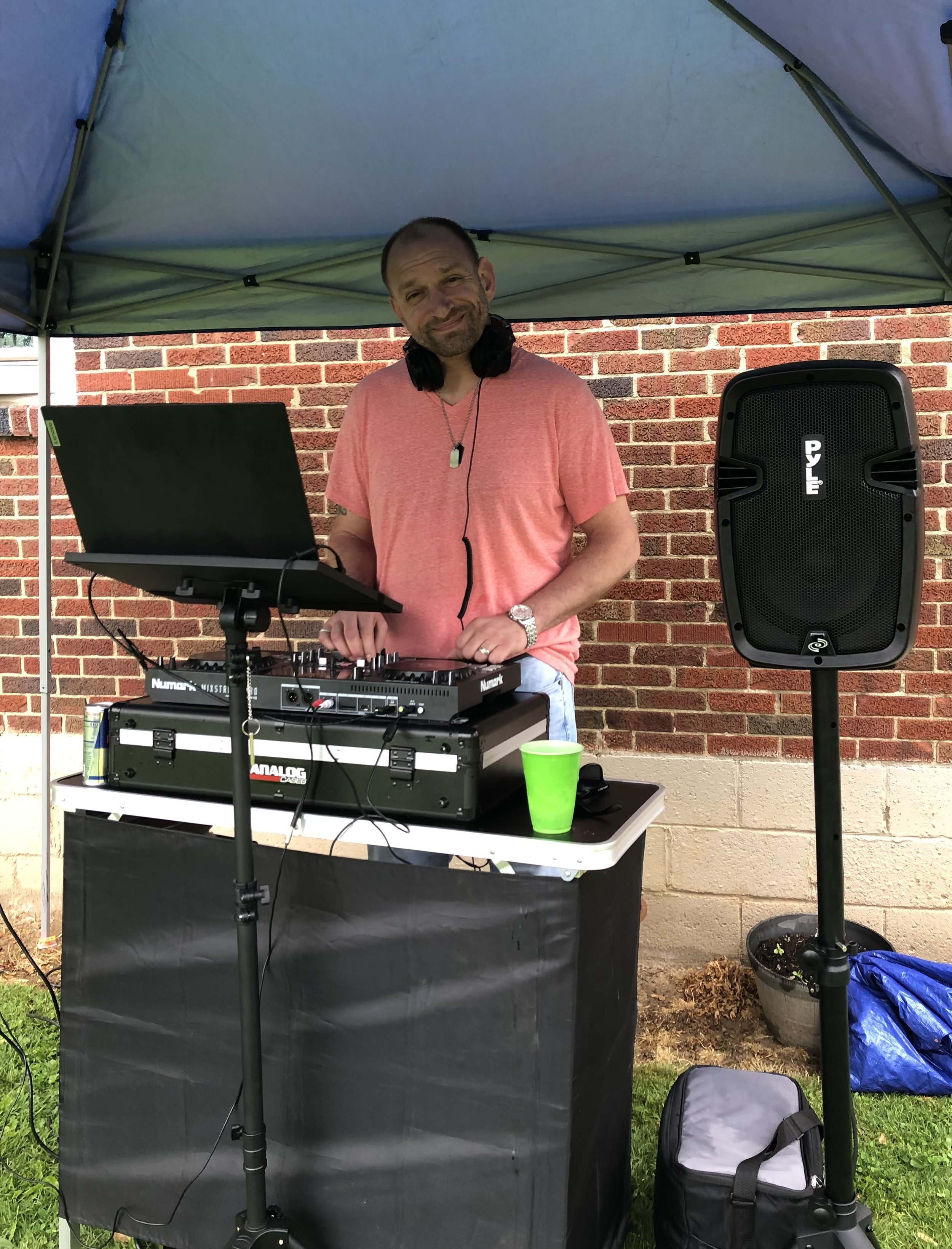 A man wearing a pink T-shirt and headphones is DJing at an outdoor event under a tent, with a brick wall background, a laptop, mixer, and speaker visible.