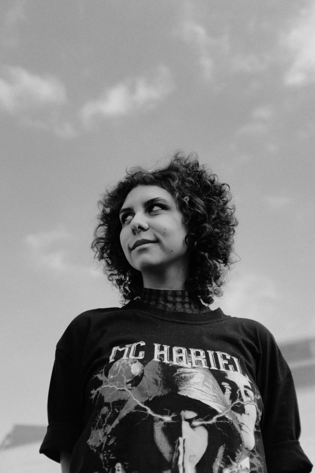 Na foto, estou de cabelos cacheados soltos, olhando para o lado esquerdo. Tenho piercing no septo, estou sorrindo sem mostrar os dentes, com uma camiseta preta do MC Hariel