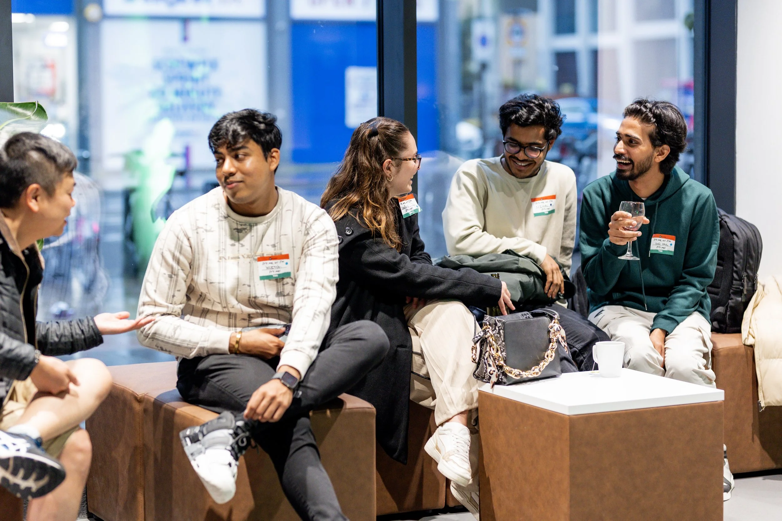 Group of people sitting and chatting in a modern indoor setting with name tags, one holding a drink, and bags nearby.