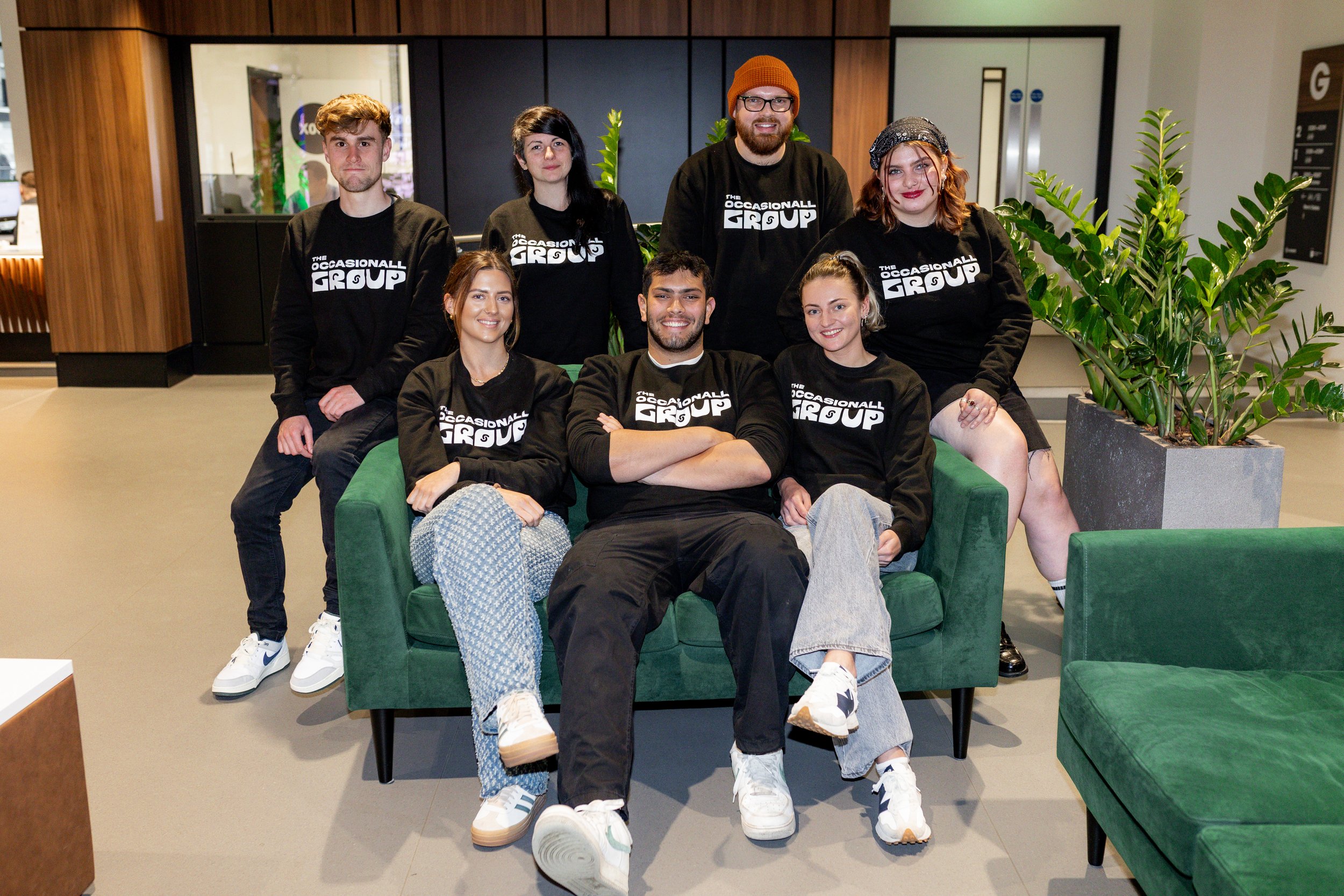 Group of seven people wearing matching black "The Occasional Group" sweatshirts, sitting and standing in an indoor environment with green couches and potted plants.