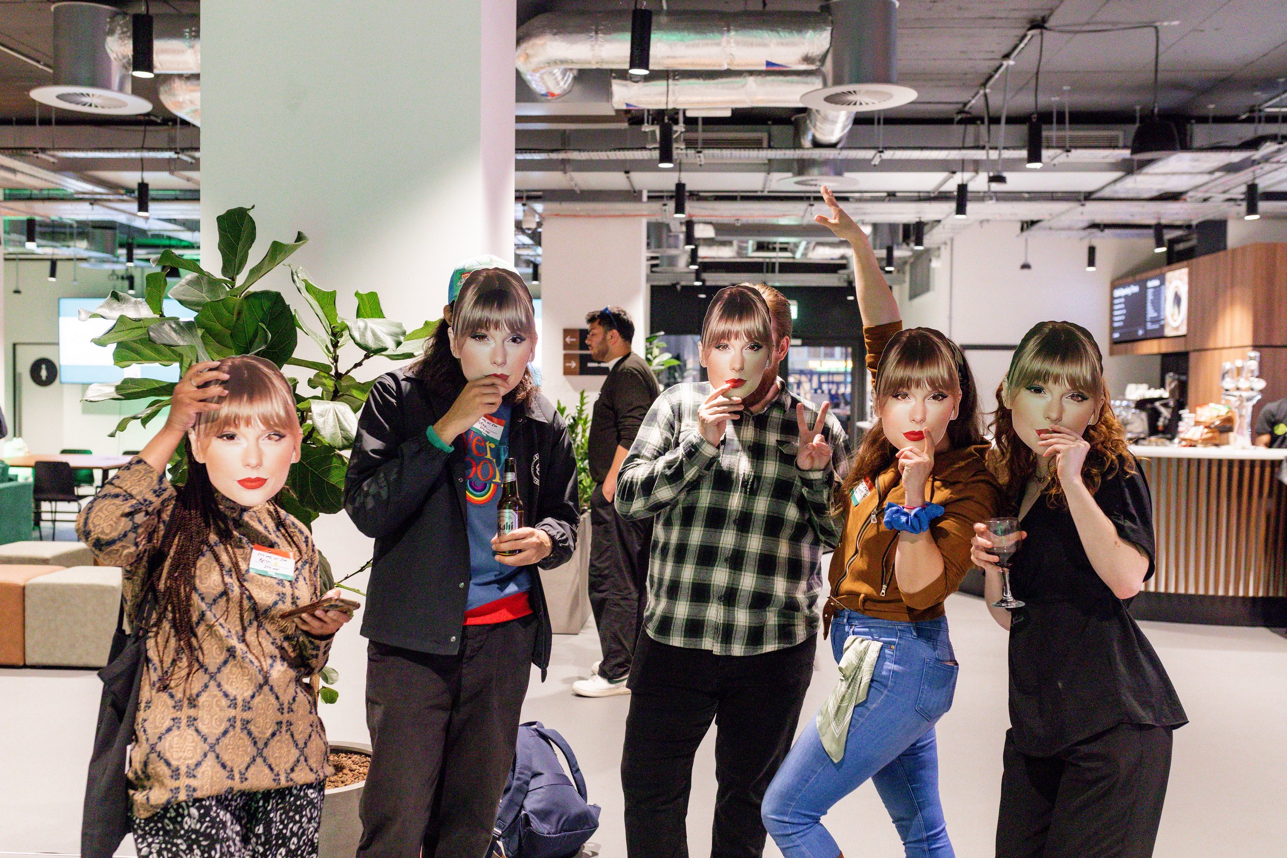 A group of five people indoors wearing identical celebrity face masks, holding drinks. The setting appears to be a modern, casual space with plants and a bar area in the background.