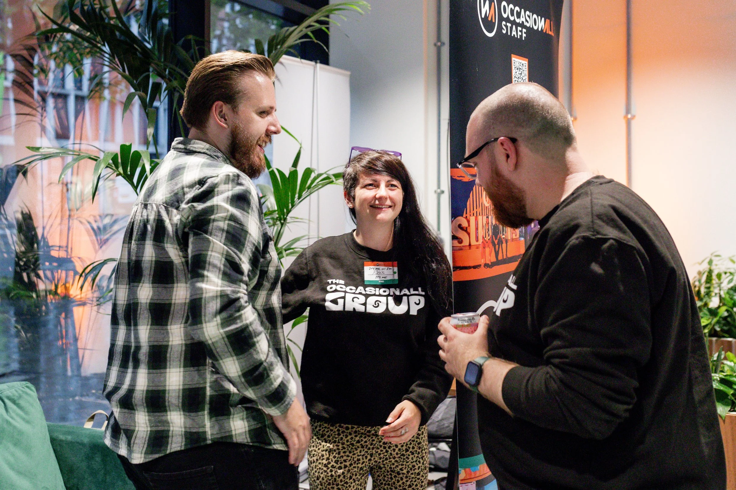 Three people at an indoor gathering, one wearing a black occasional group sweatshirt, another in a plaid shirt, and the third holding a beverage and wearing glasses. They appear to be socializing near a branded banner.