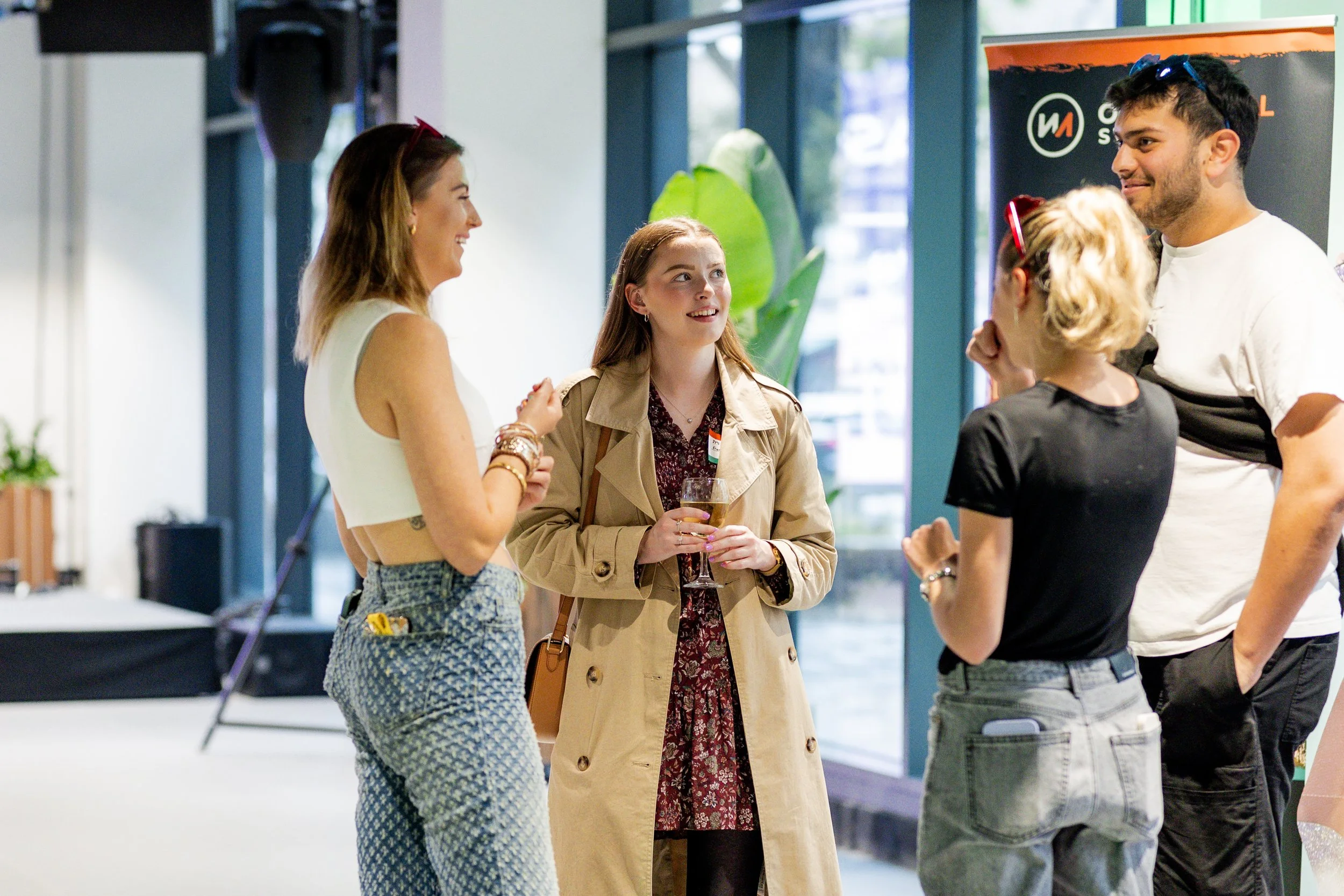 A group of four people having a casual conversation indoors. They are standing and smiling, with one person holding a drink. The setting appears to be a modern, well-lit space with large windows and greenery.