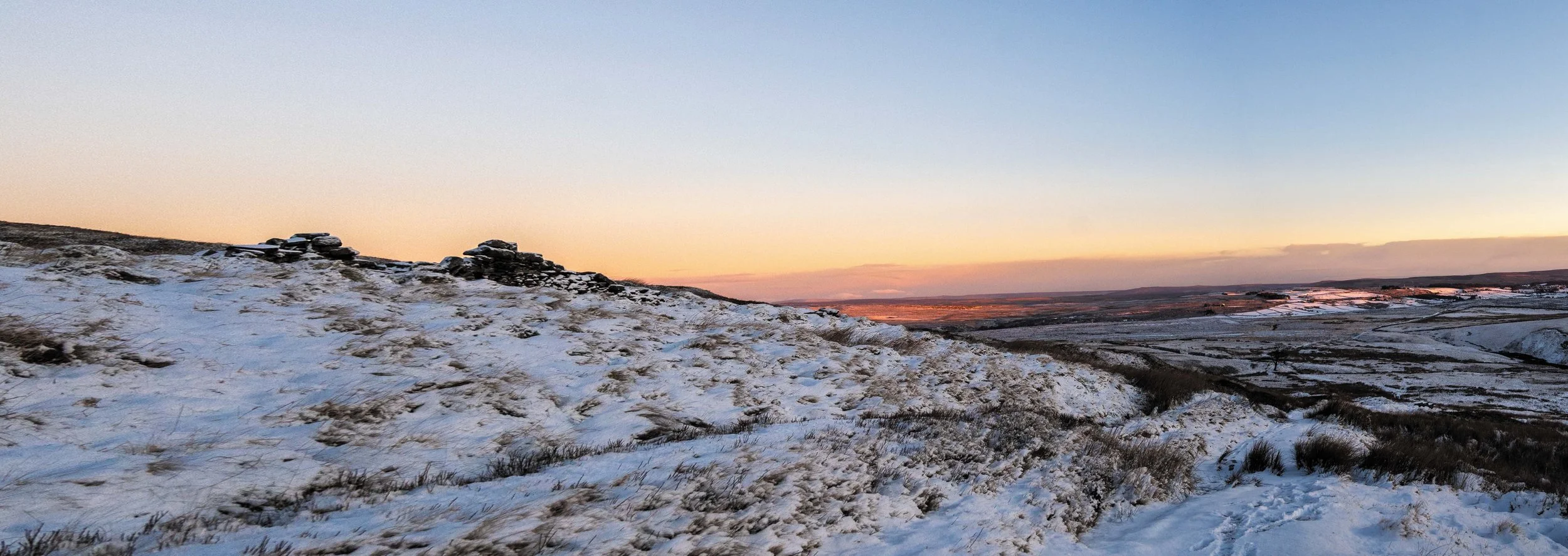 Sneeuw bedekte heuvels en velden bij zonsondergang, met een heuvel met stenen en gras op de voorgrond en een uitgestrekt landschap op de achtergrond.
