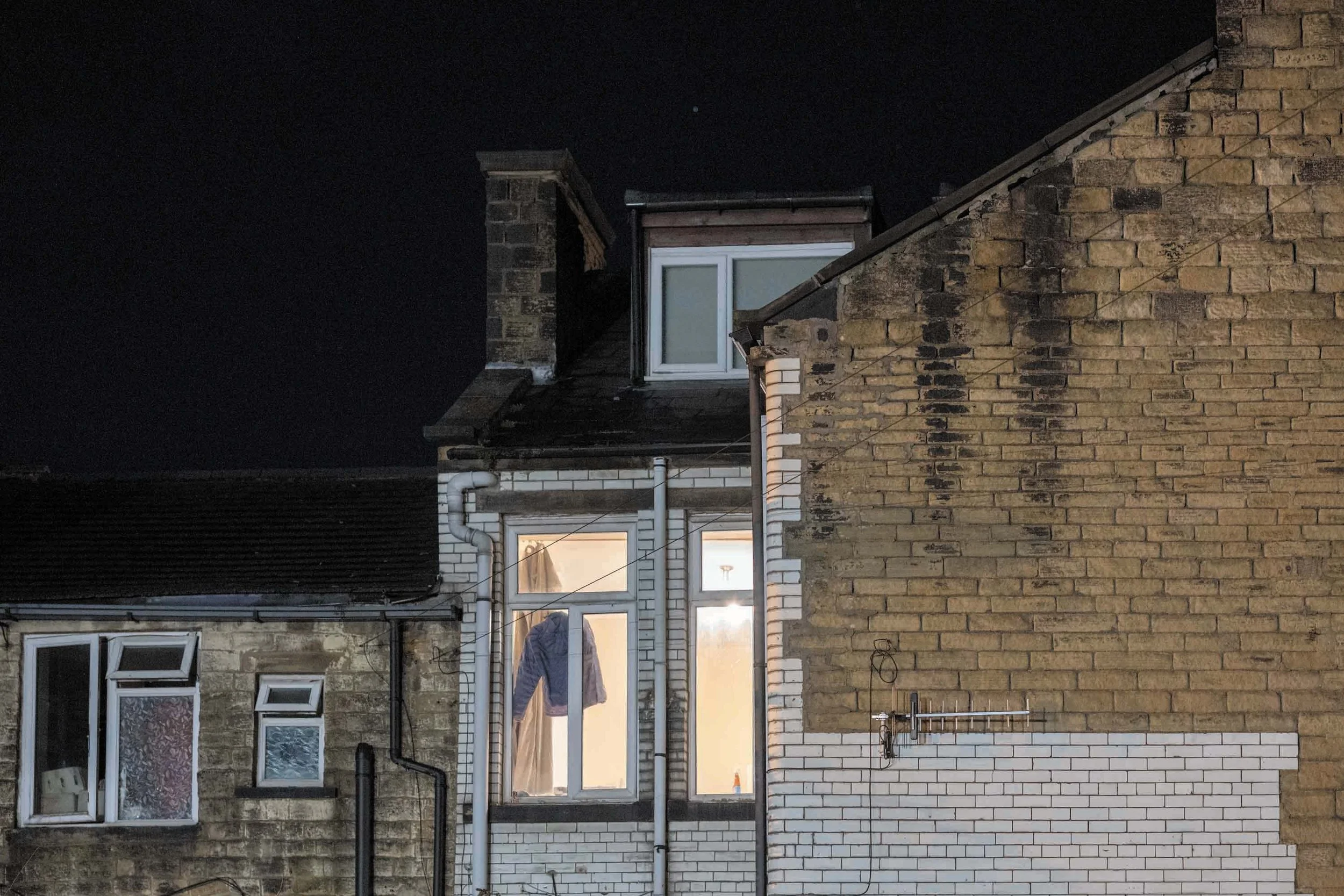 Bovenaanzicht van een rij woningen met diverse ramen en gevels, waarvan sommige openstaan en verlichte interieurs zichtbaar zijn, onder een donkere nachtelijke hemel.