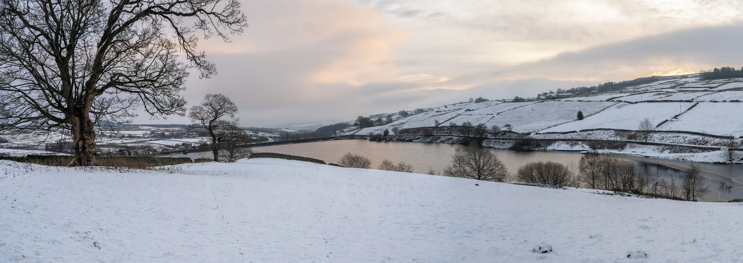 Sneeuw bedekte heuvels, een grote bladerenloze boom, een rivier, en een ondergaande zon met een bewolkte lucht in een landelijke landschap.