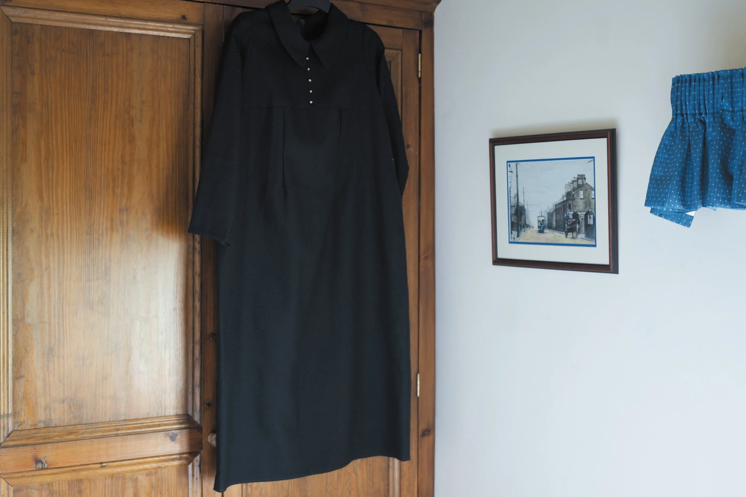 Een zwart kledingstuk, mogelijk een jurk of een lang shirt, hangt aan een houten kastdeur. Daarnaast hangt een blauwe short met witte stippen, rechts op de muur, naast een ingelijste foto van een stad met gebouwen en een tram.