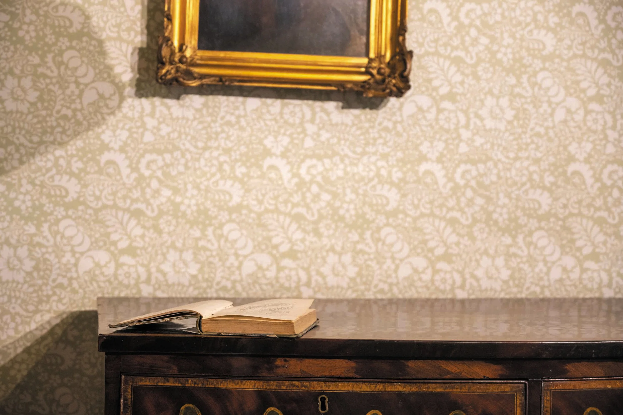 Open boek ligt op een houten kastje met een lade, achtergrond met sierlijk patroon op de muur.