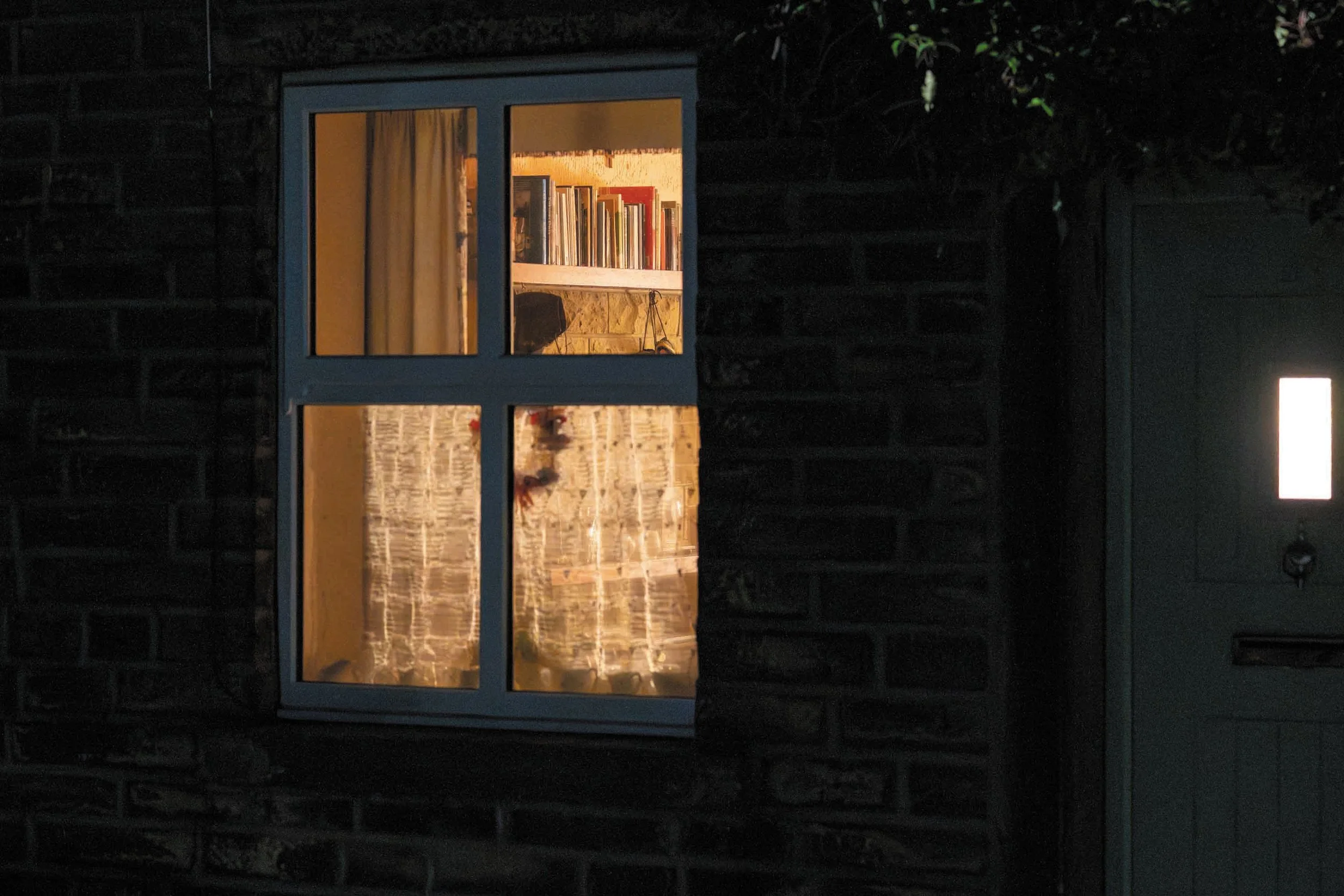 Gezellige boekenkast met boeken die binnen in een warm verlichte kamer staan, gezien door een raam in een donkere buitenruimte.