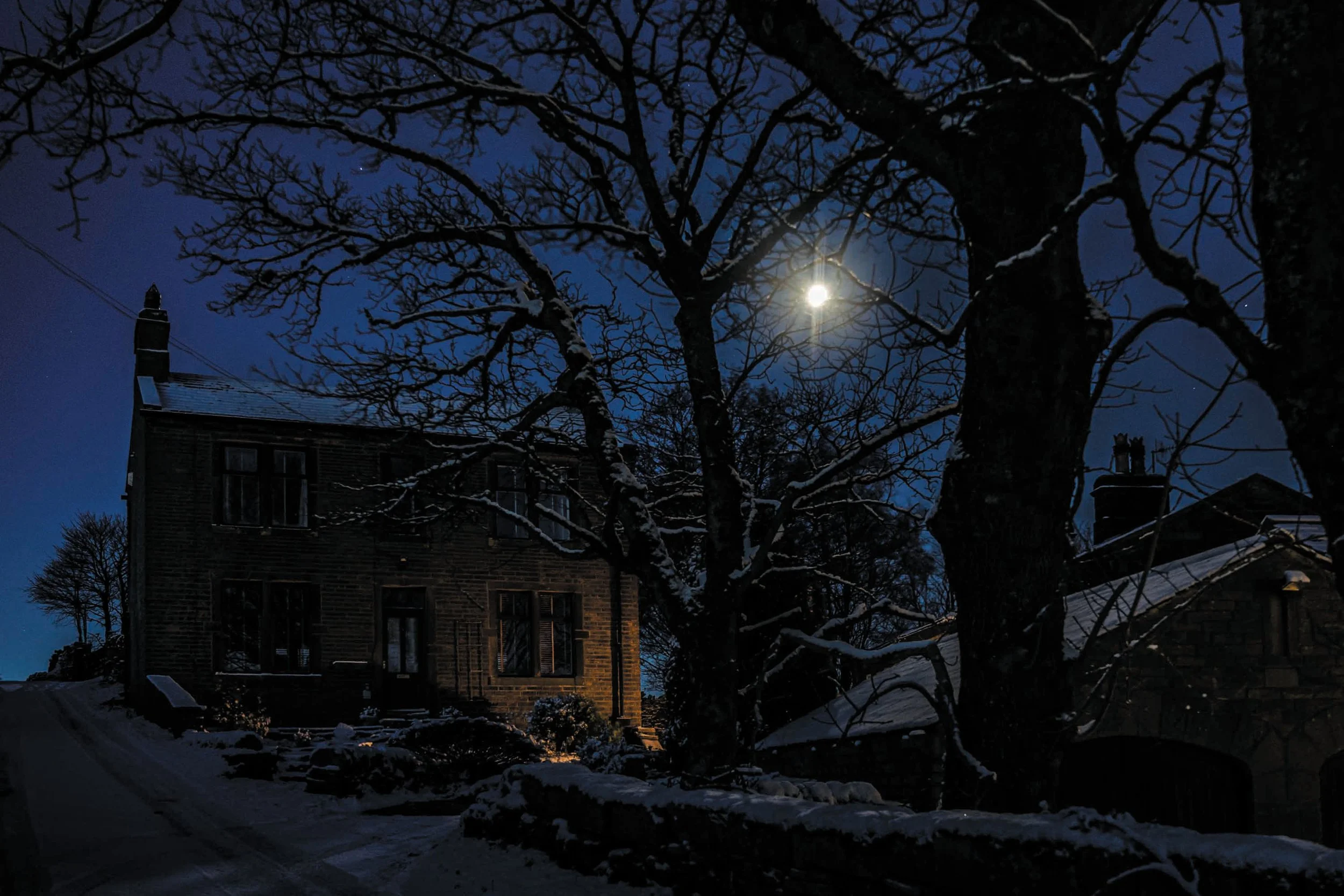 Een donker winteravond met een heldere maan die door de takken van een boom schijnt, een oude stenen woning met verlichte ramen en een sneeuwbedekte tuin.